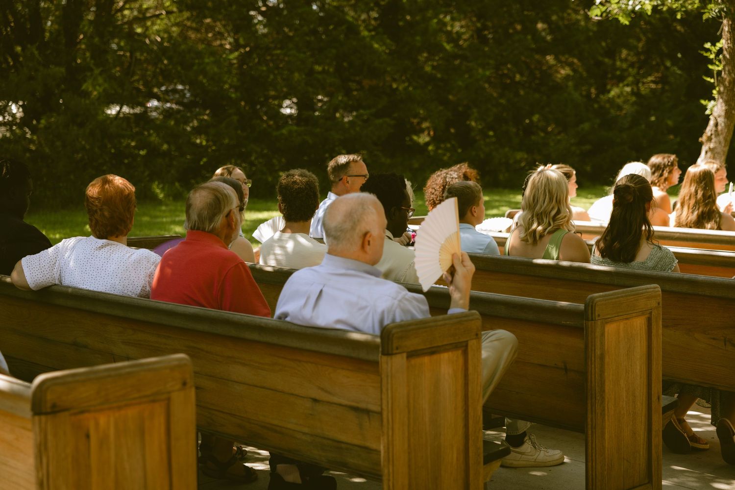 Wedding guests seated on wooden pews during an outdoor ceremony surrounded by trees in warm sunlight.