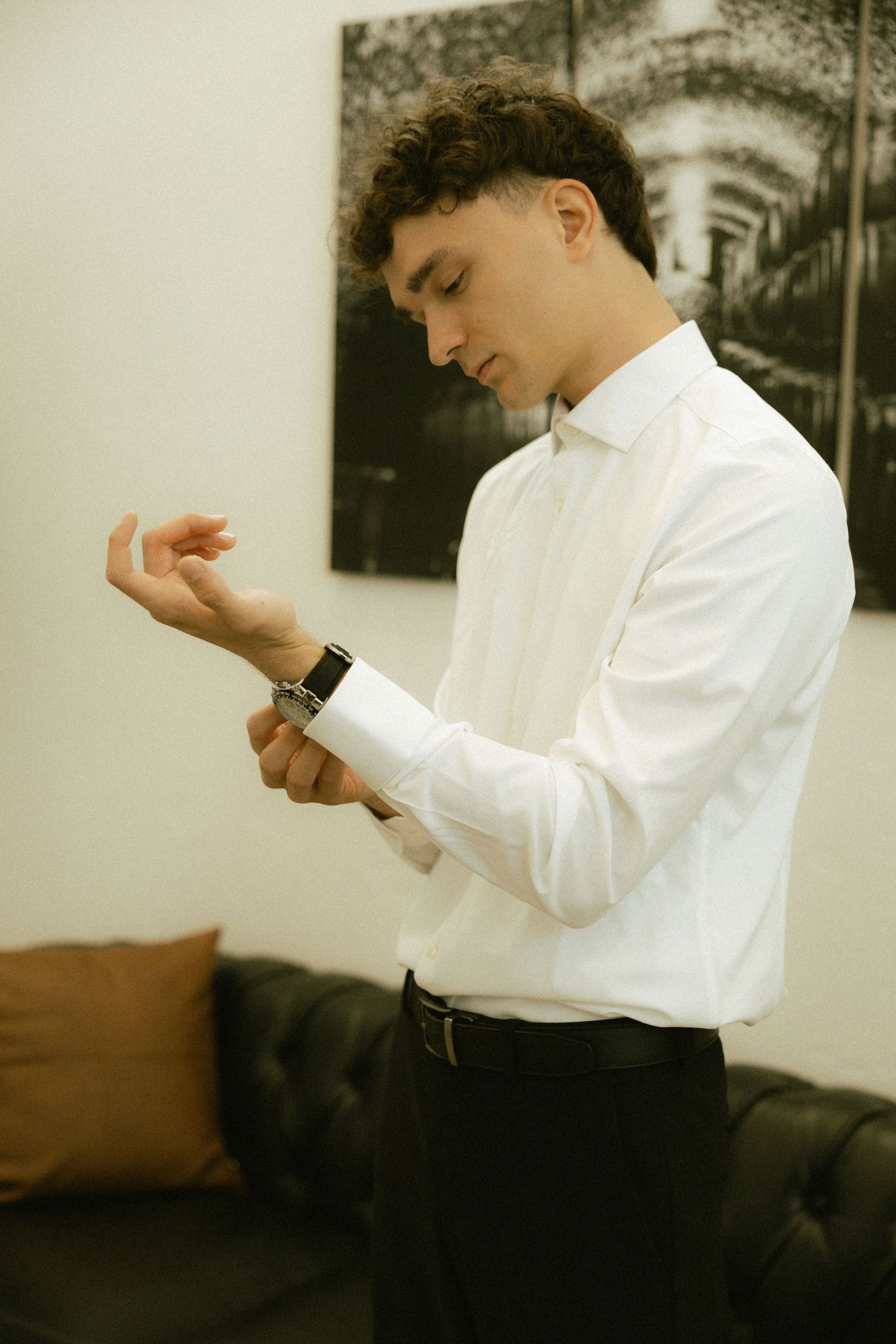 Young man in white dress shirt adjusting his watch cufflink indoors near a black leather sofa.