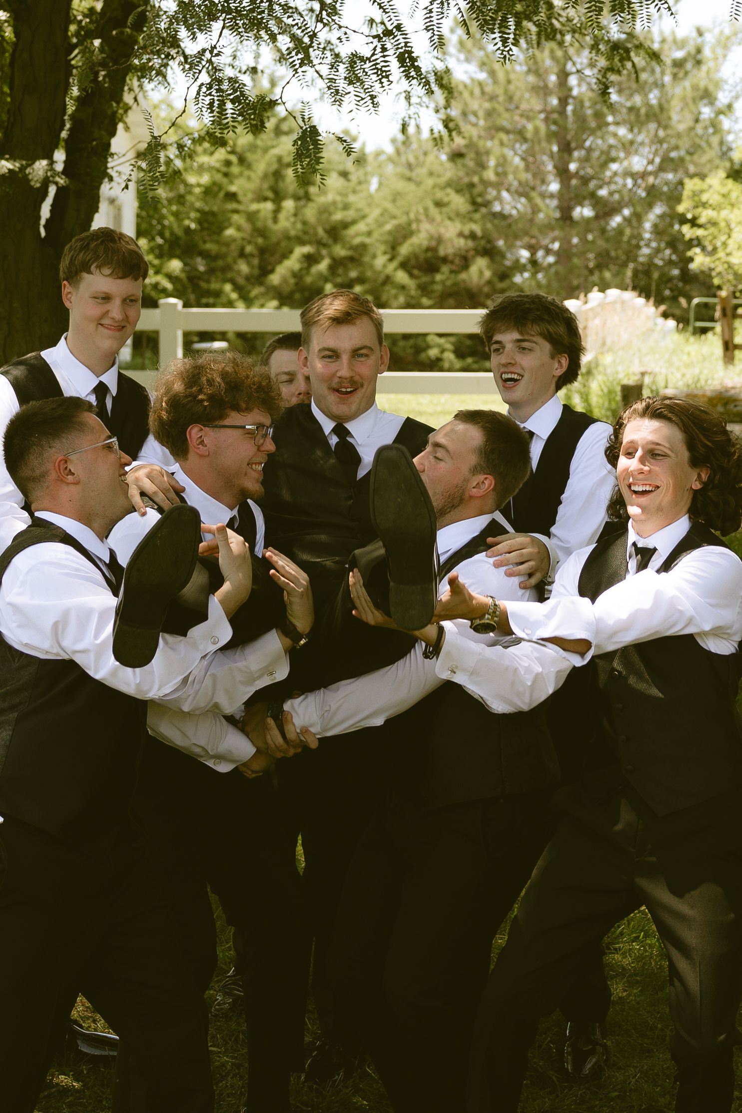 Groom surrounded by laughing groomsmen in black vests and white shirts in outdoor wedding photo.