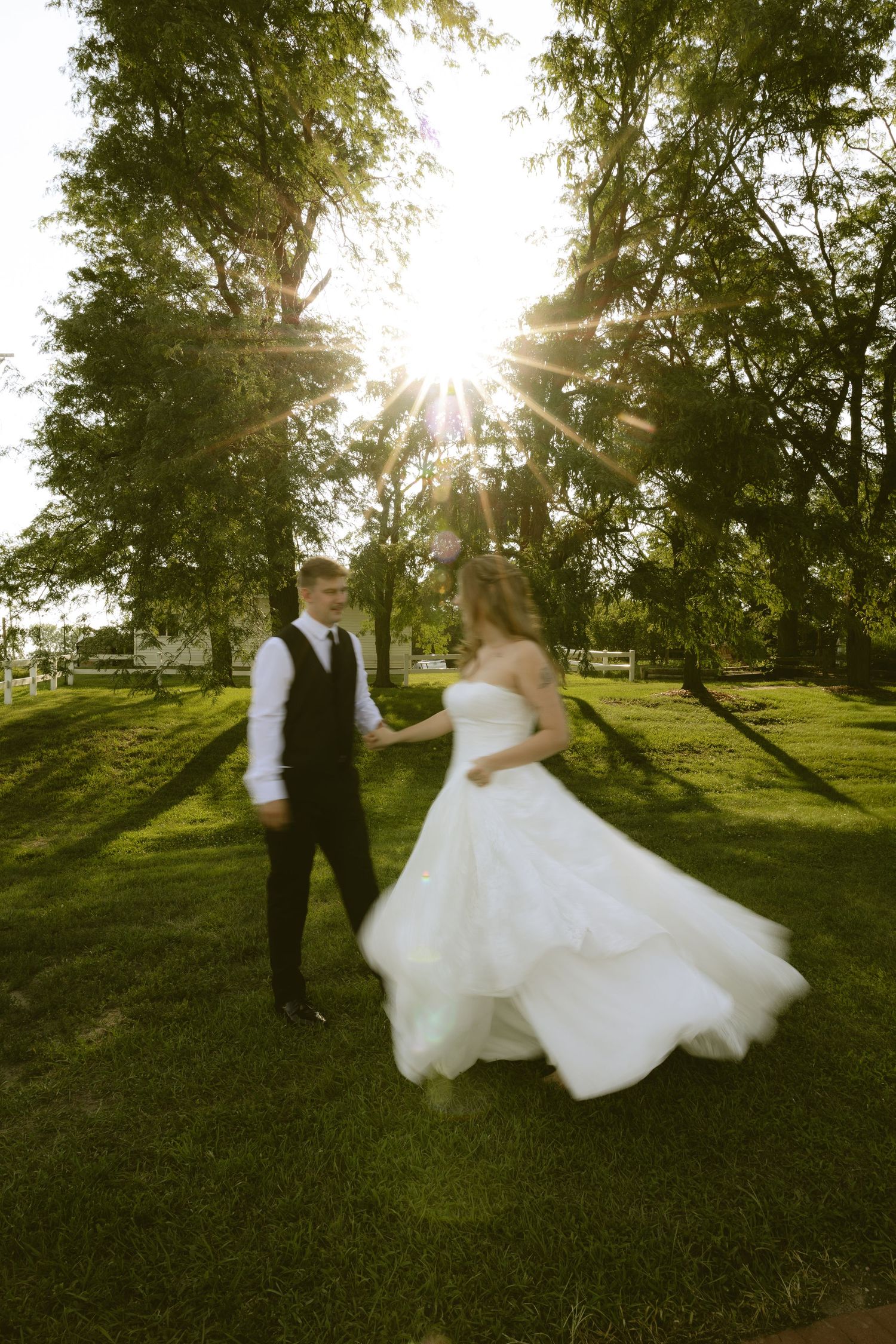 Bride and groom hold hands dancing in sunlit garden, bride's white ball gown flowing in motion.