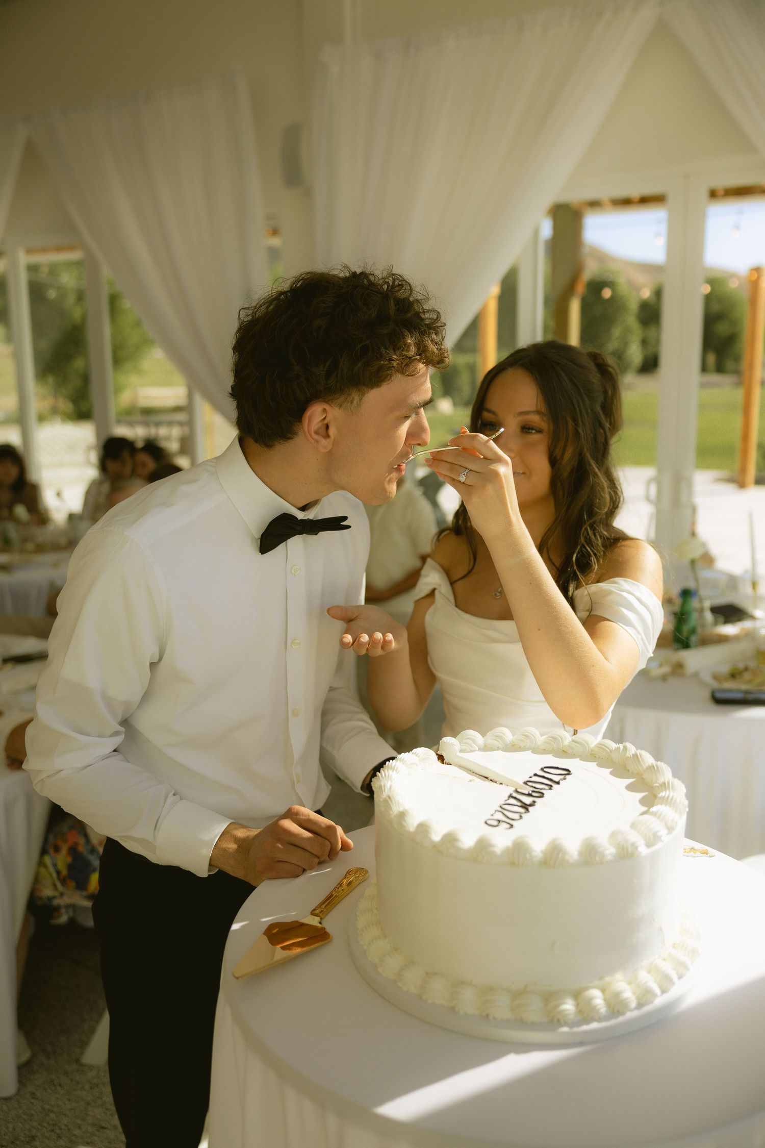 Bride and groom cutting white wedding cake at outdoor reception under elegant white draped tent.
