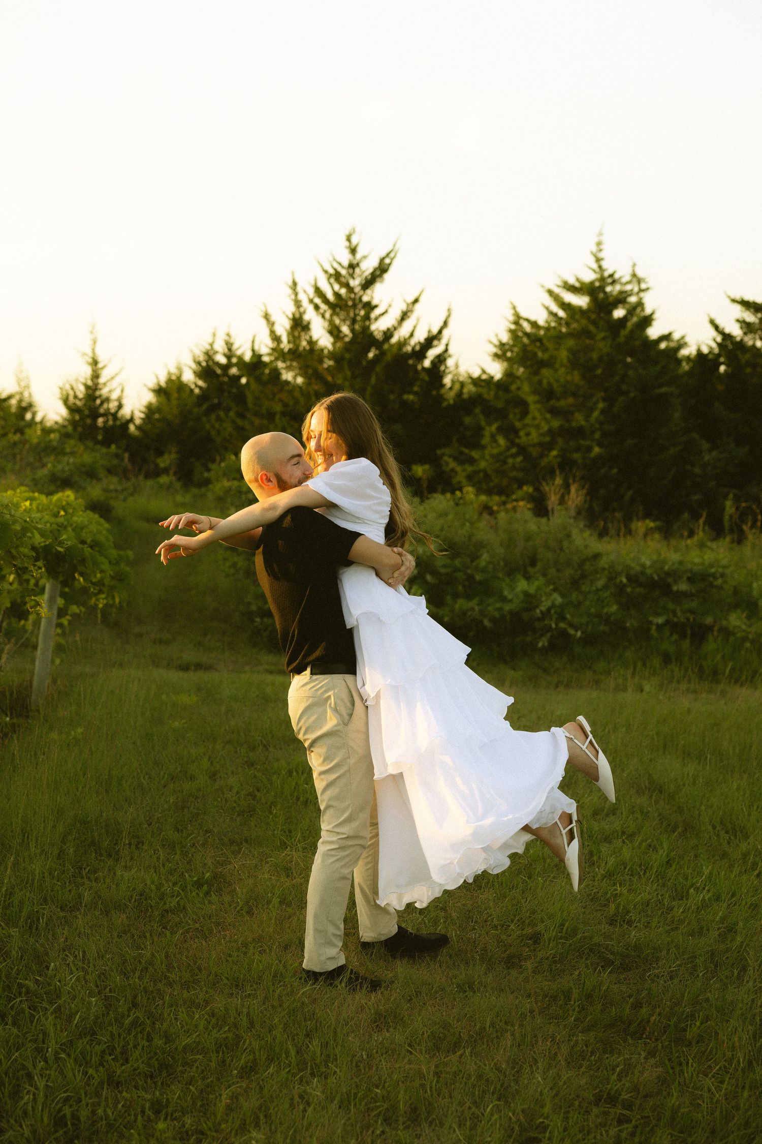 A man lifts a woman in a white dress outdoors at golden hour, surrounded by lush green trees and grass.