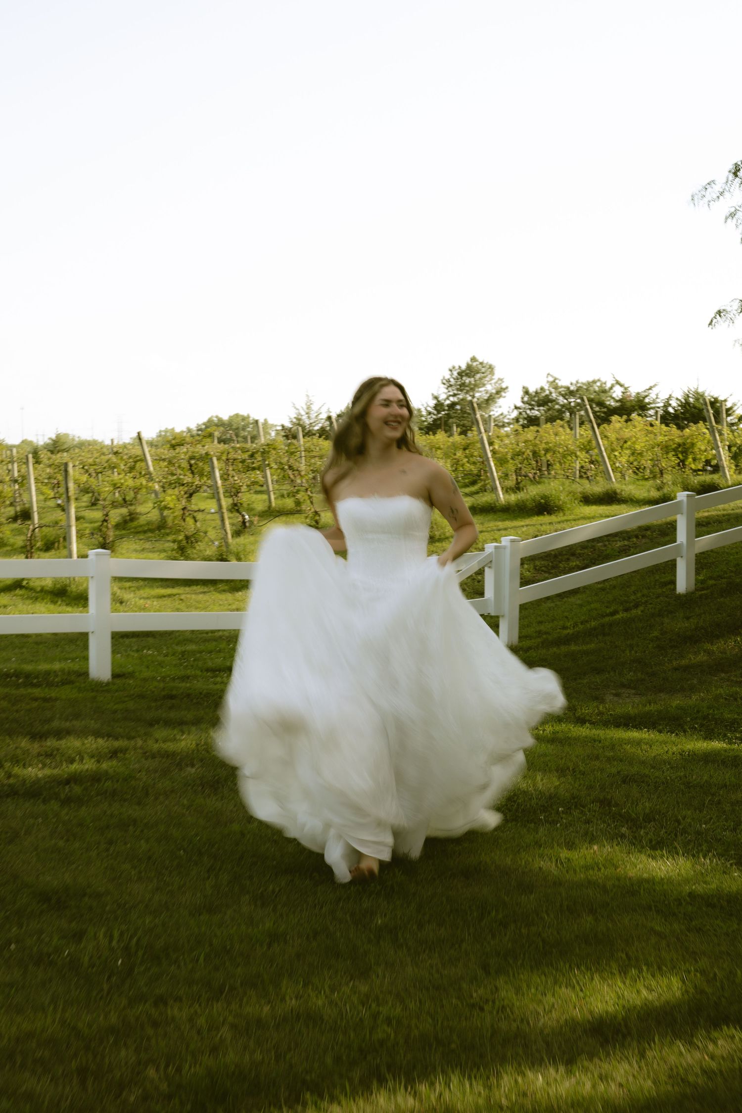 Smiling bride in a flowing white strapless tulle gown twirling on green grass near a white fence and vineyard.