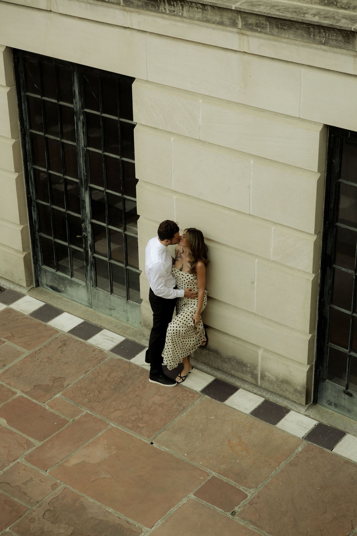 Two people share an intimate moment against a white building wall with windows.