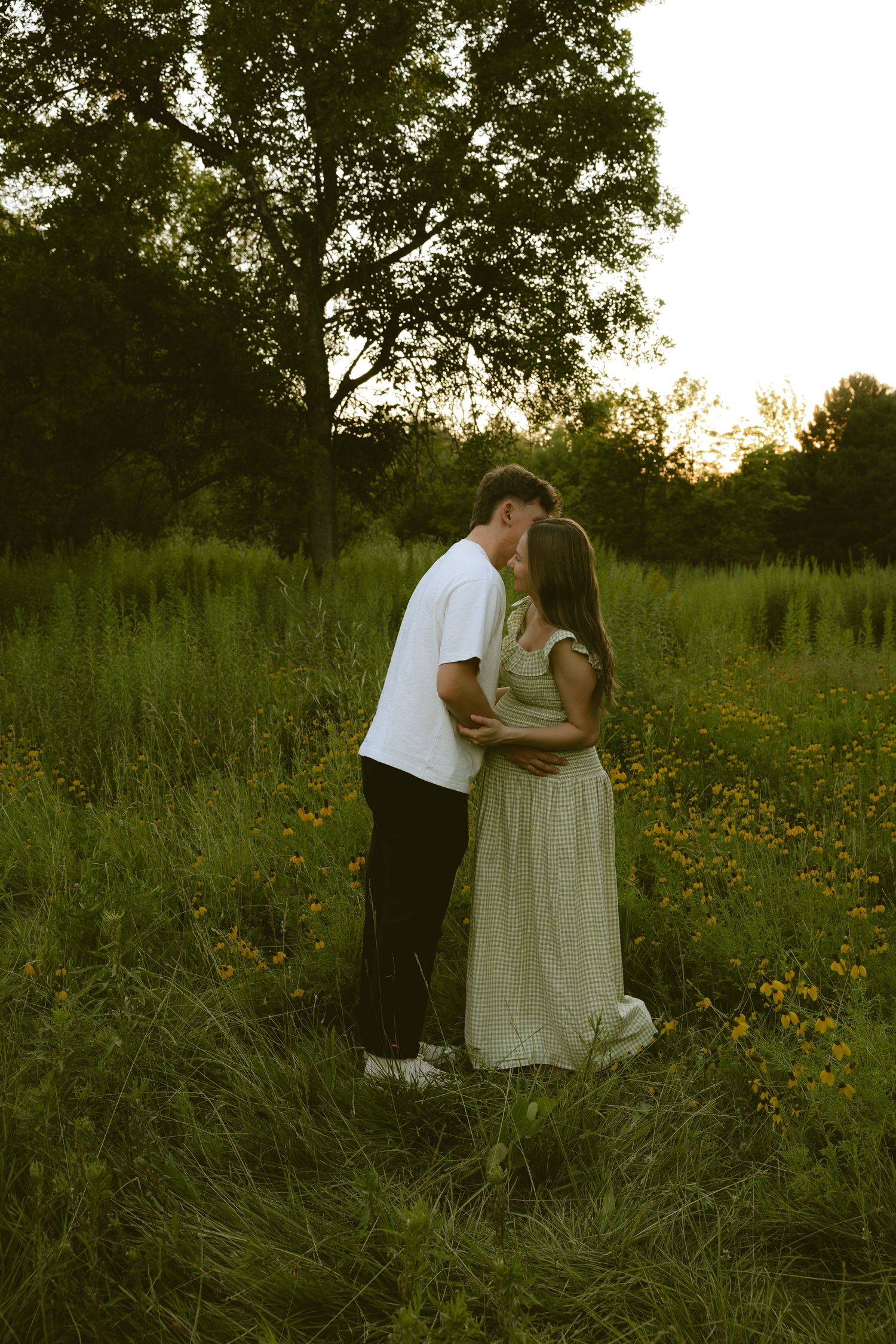 Couple sharing a romantic kiss in a wildflower meadow at golden hour, surrounded by lush green grass and trees.