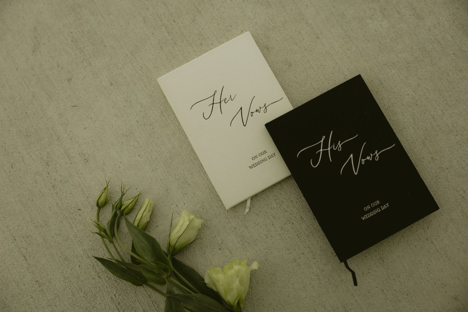 His and Her Vows booklets in black and white with elegant script lettering, beside white roses on a marble surface.