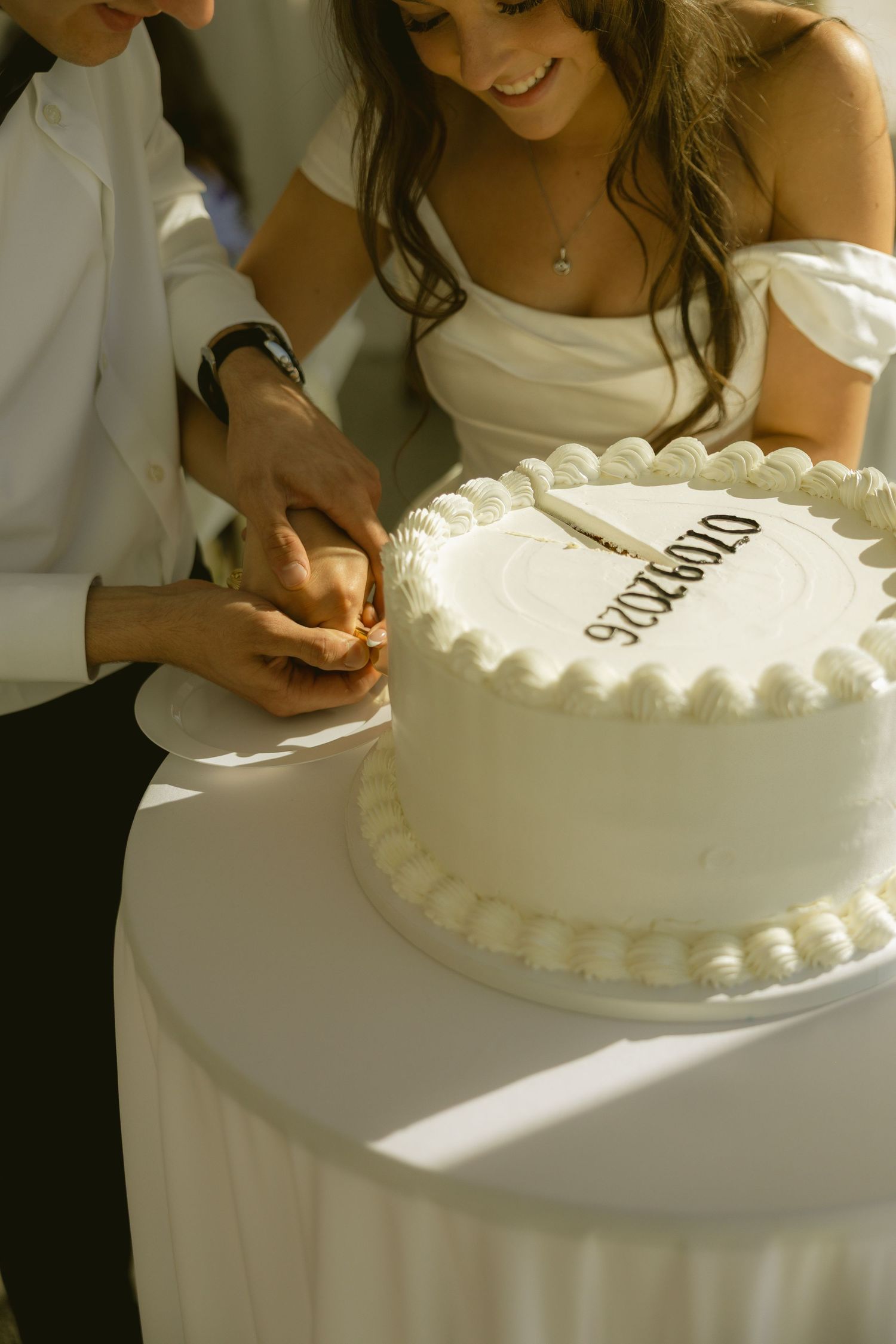 Bride and groom cutting white wedding cake with date inscription at outdoor reception ceremony.