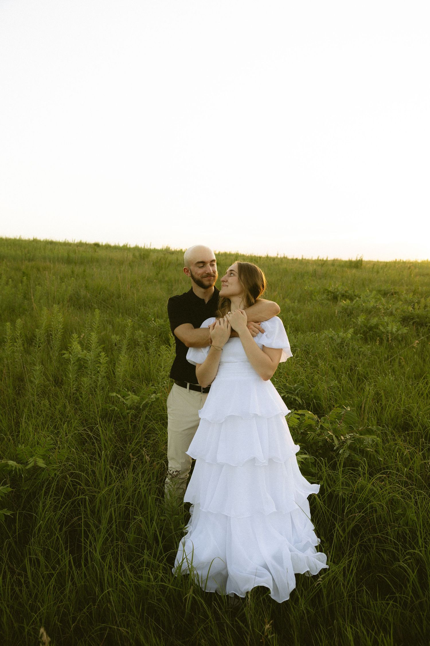 Couple embracing in green field at sunset, woman in white tiered ruffle dress, man in black shirt.