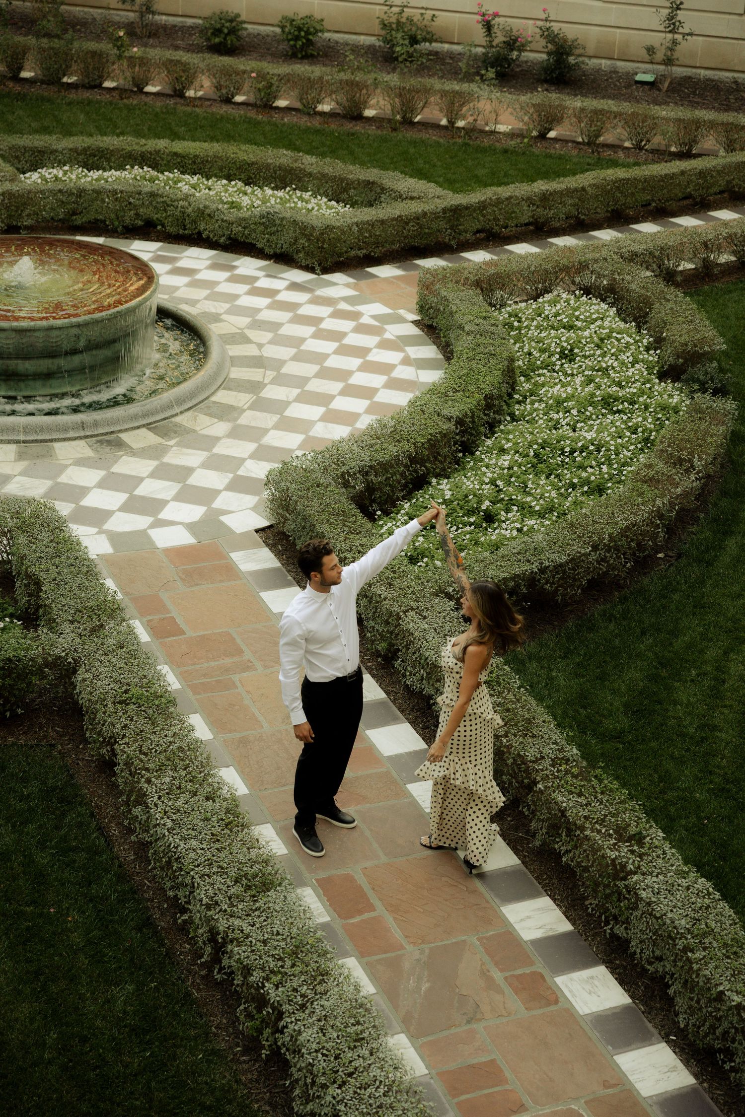 Two people dancing on a geometric patterned garden path surrounded by manicured hedges and a decorative fountain.