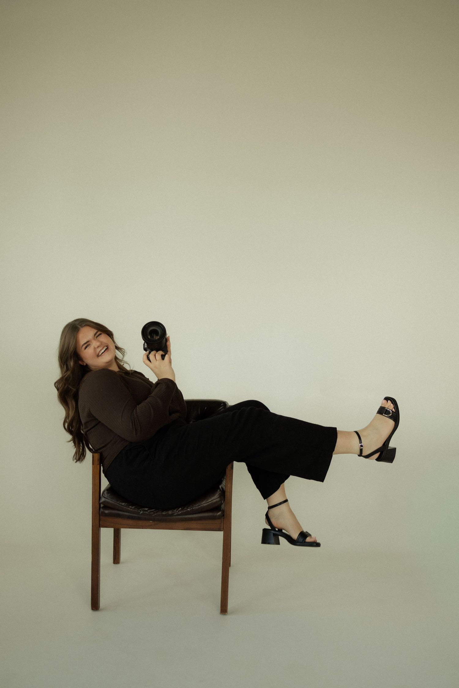 Woman in black outfit sitting relaxed on wooden chair, smiling and holding a camera against light background.