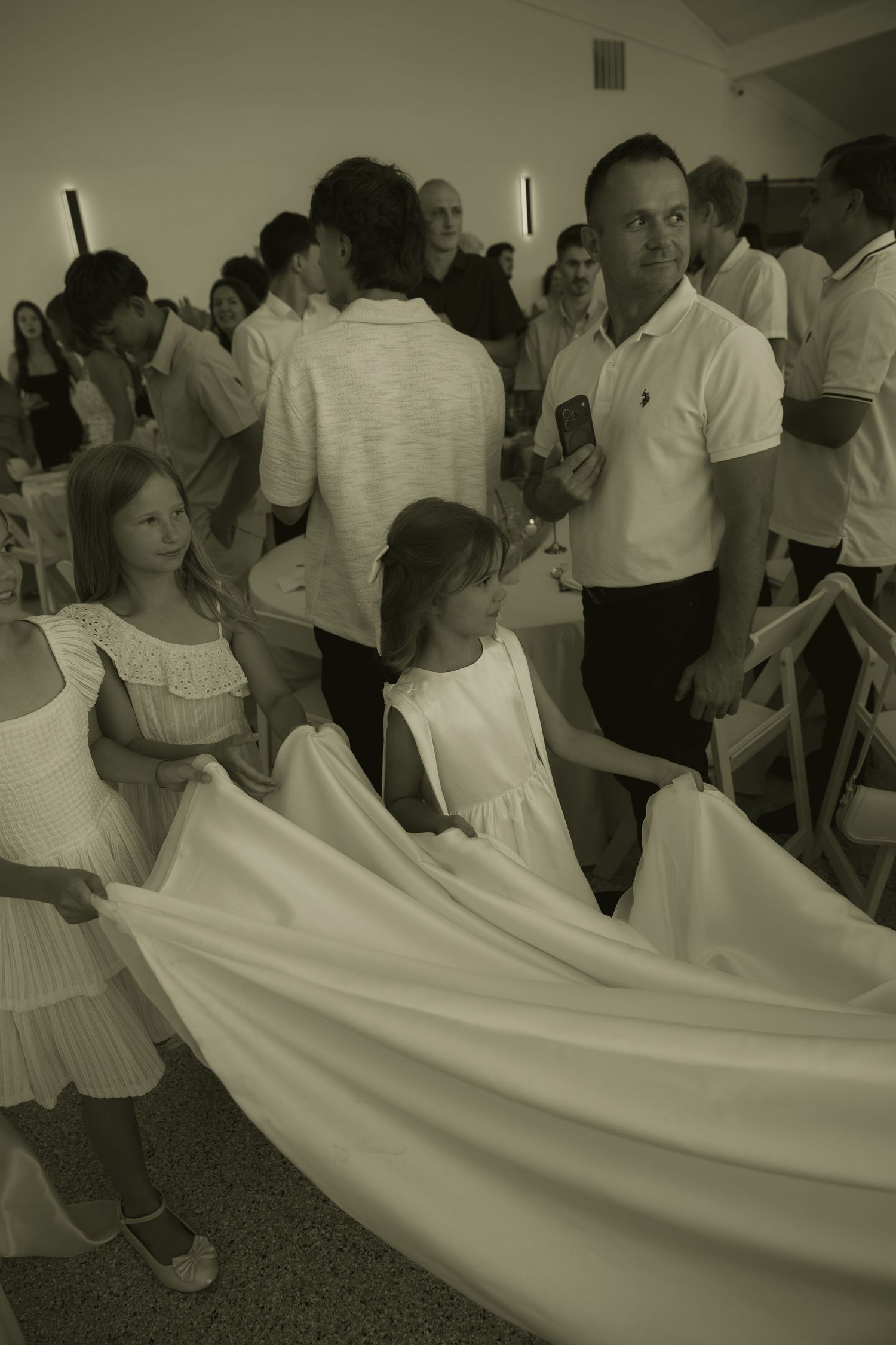 Two young girls in white dresses hold a bridal gown train at a wedding ceremony indoors.