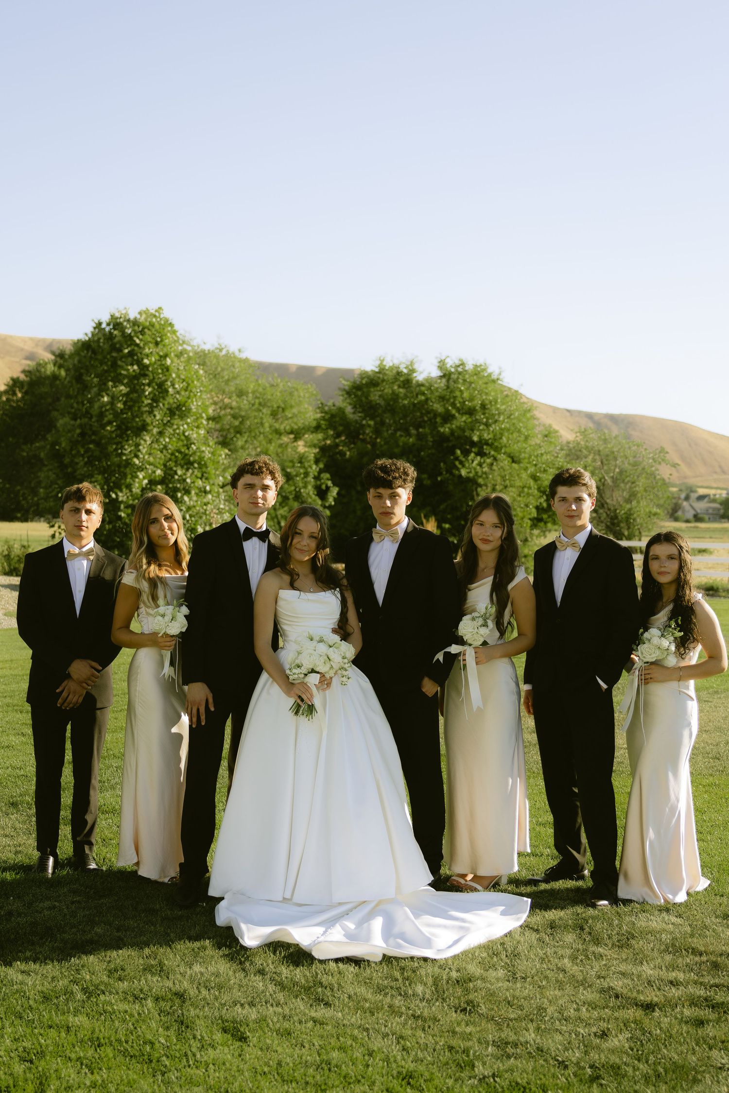 Wedding party posing outdoors in black tuxedos and white gowns with rolling hills behind.