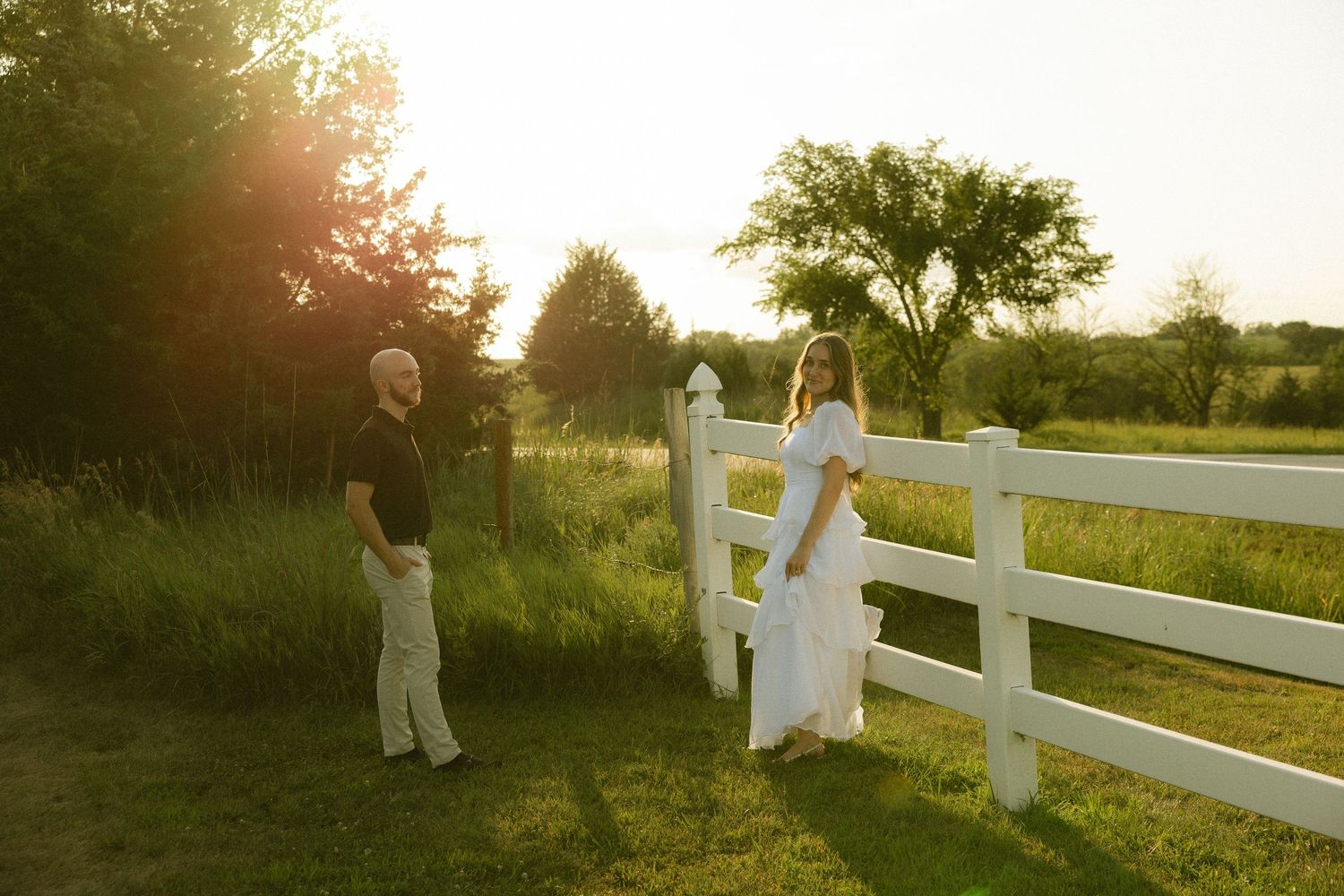Couple standing by white fence at golden hour sunset in lush green countryside setting.