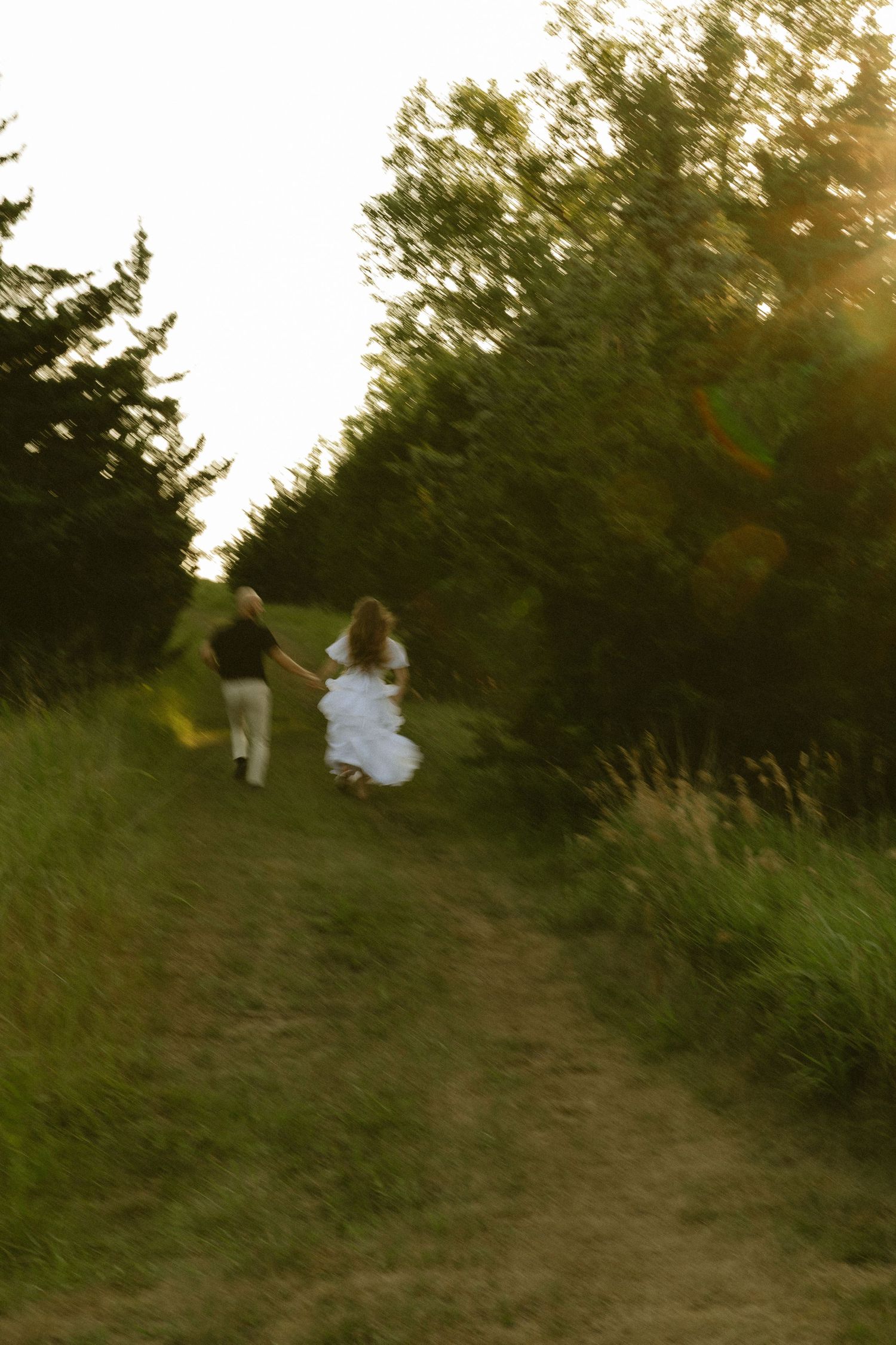 A couple in wedding attire runs hand-in-hand down a sunlit path lined with lush green trees at golden hour.