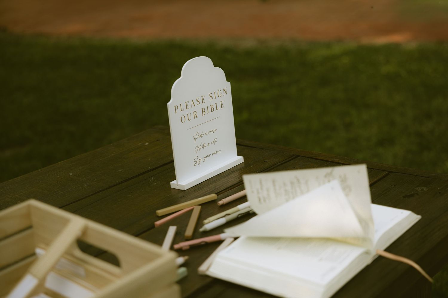 Wedding guest book table with a sign reading Please Sign Our Bible, open book, and wooden crate with pens.