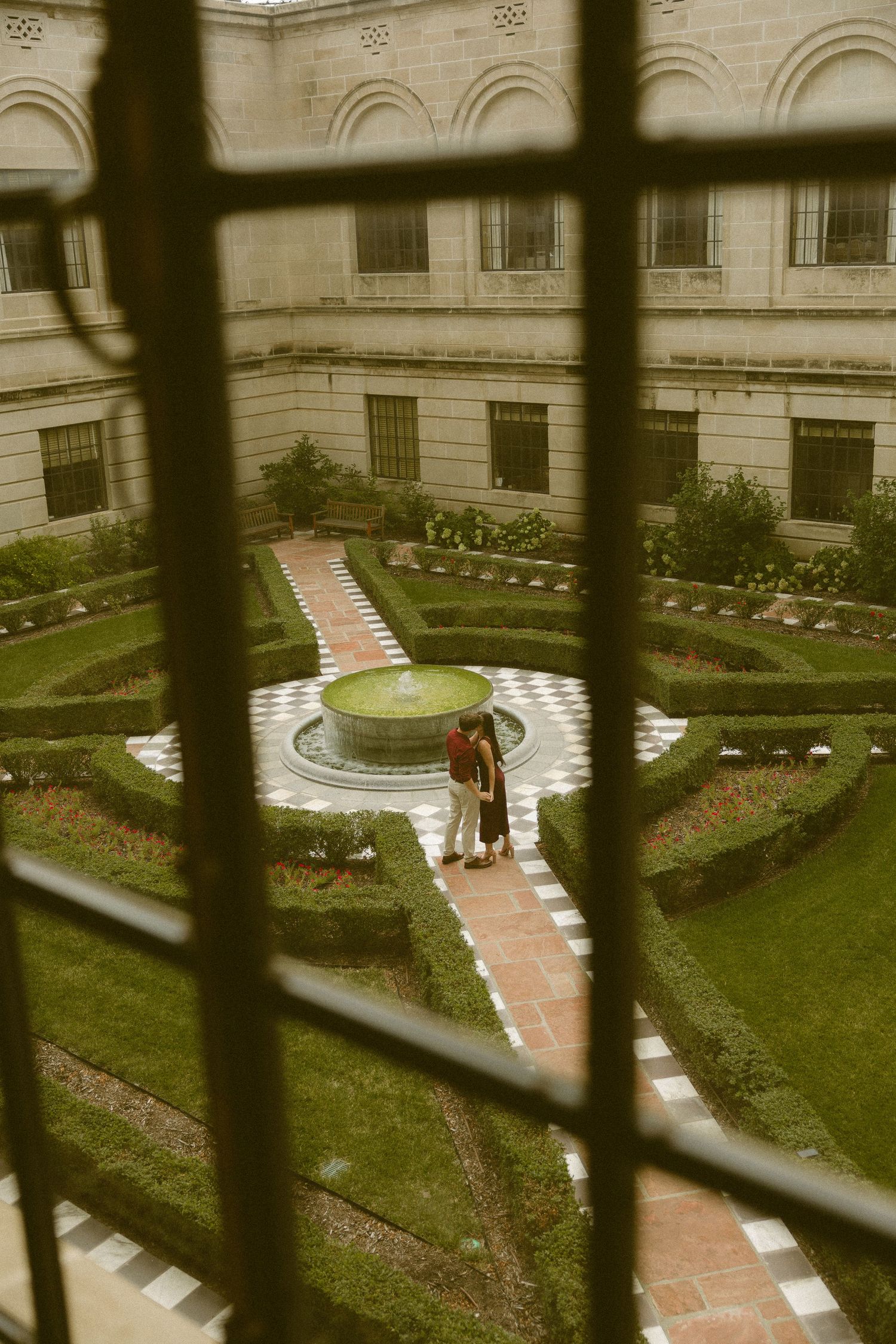 Couple embracing near a fountain in a formal garden courtyard, viewed through iron bars of a window.