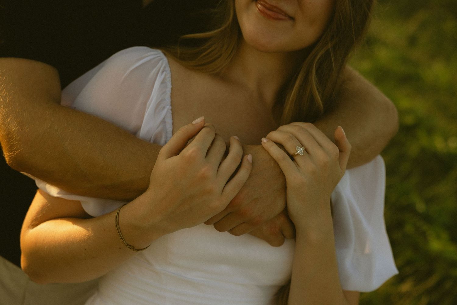 Couple embracing outdoors, woman in white off-shoulder top wearing engagement ring, man's arms wrapped around her.