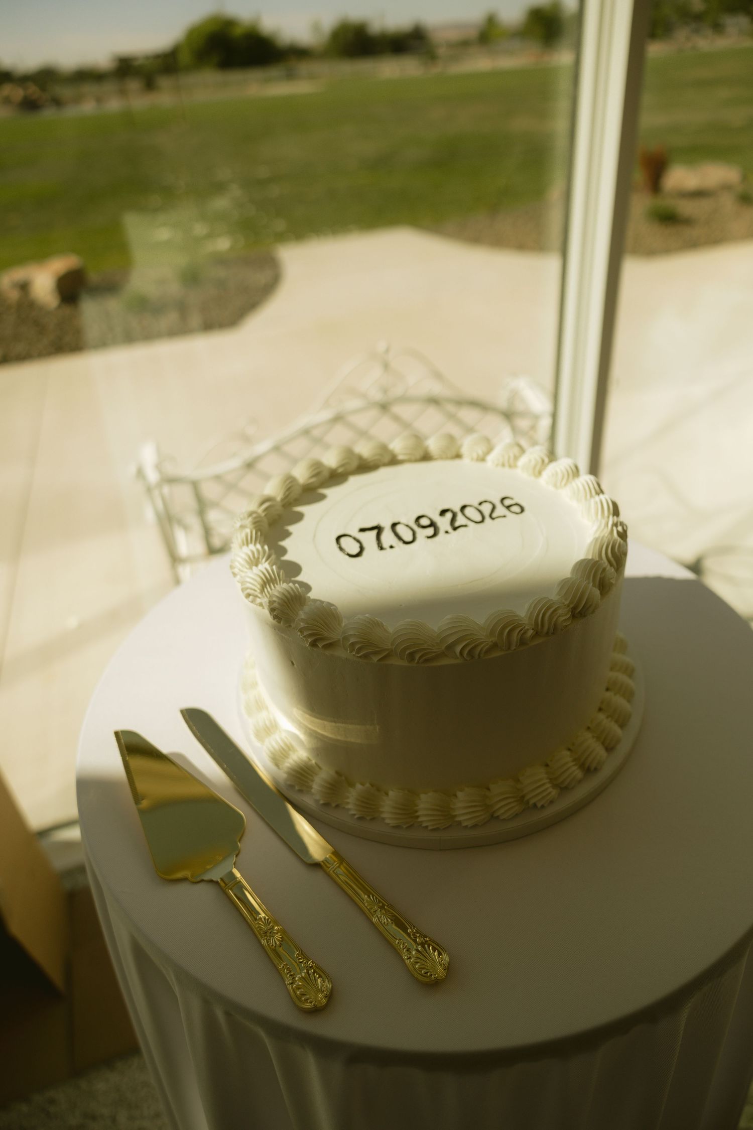 White wedding cake with date 07.09.2026 displayed on a round table with gold cake serving set near a window.