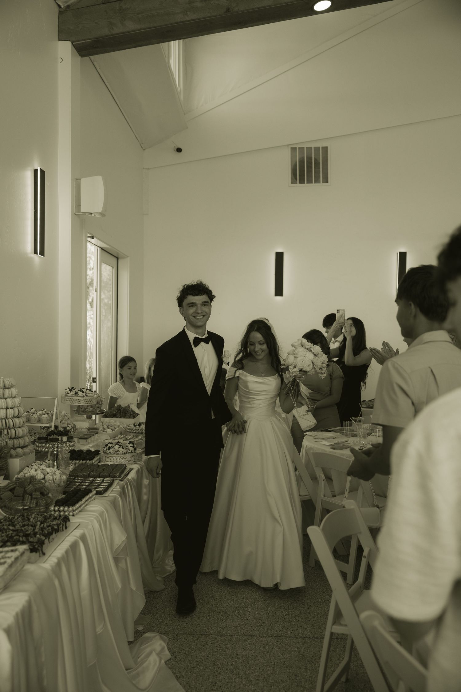 Bride and groom walk hand-in-hand at their wedding reception in a bright venue, guests applauding around decorated tables.