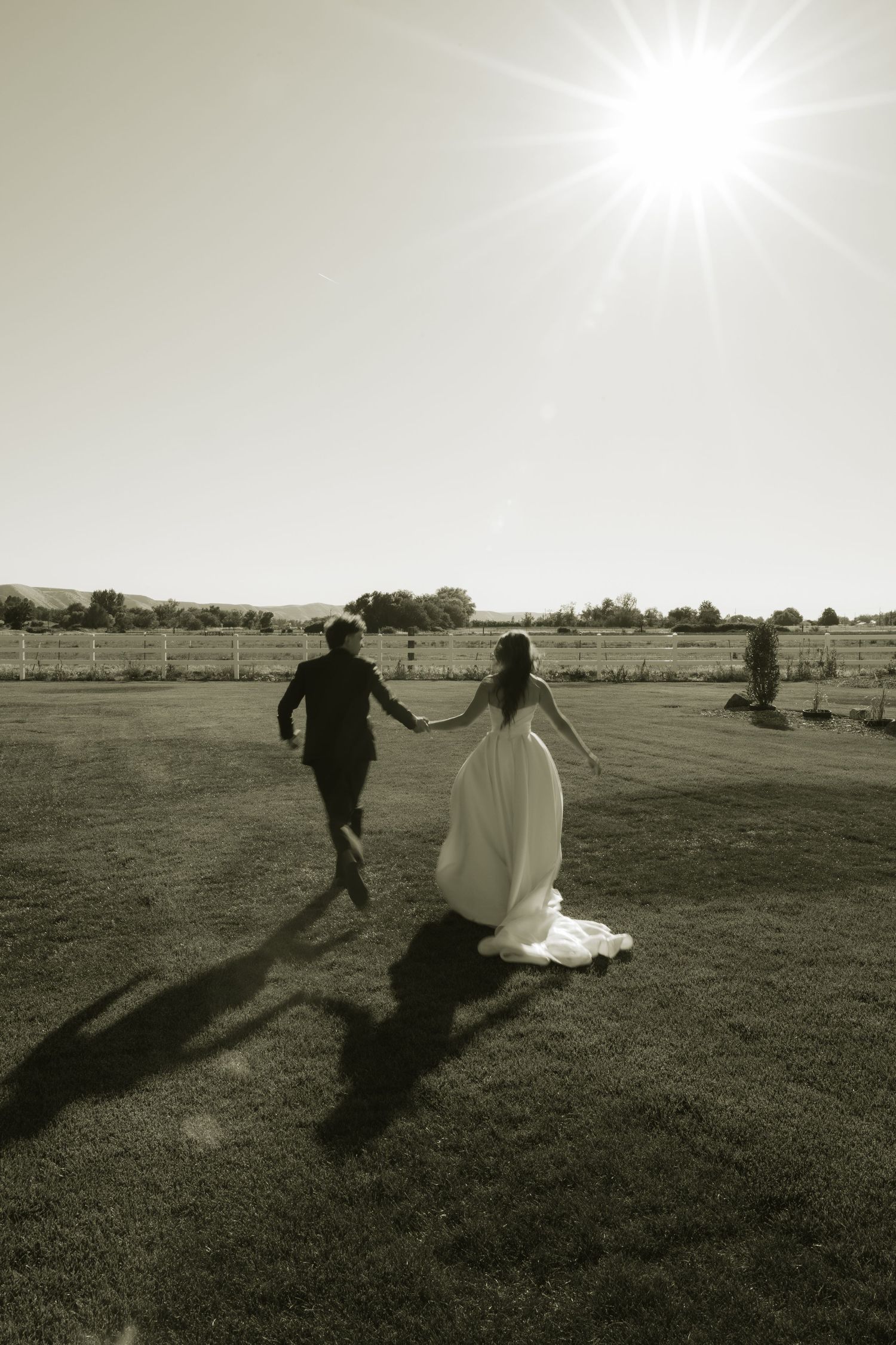 Black and white photo of bride and groom running hand-in-hand across sunny farm field.