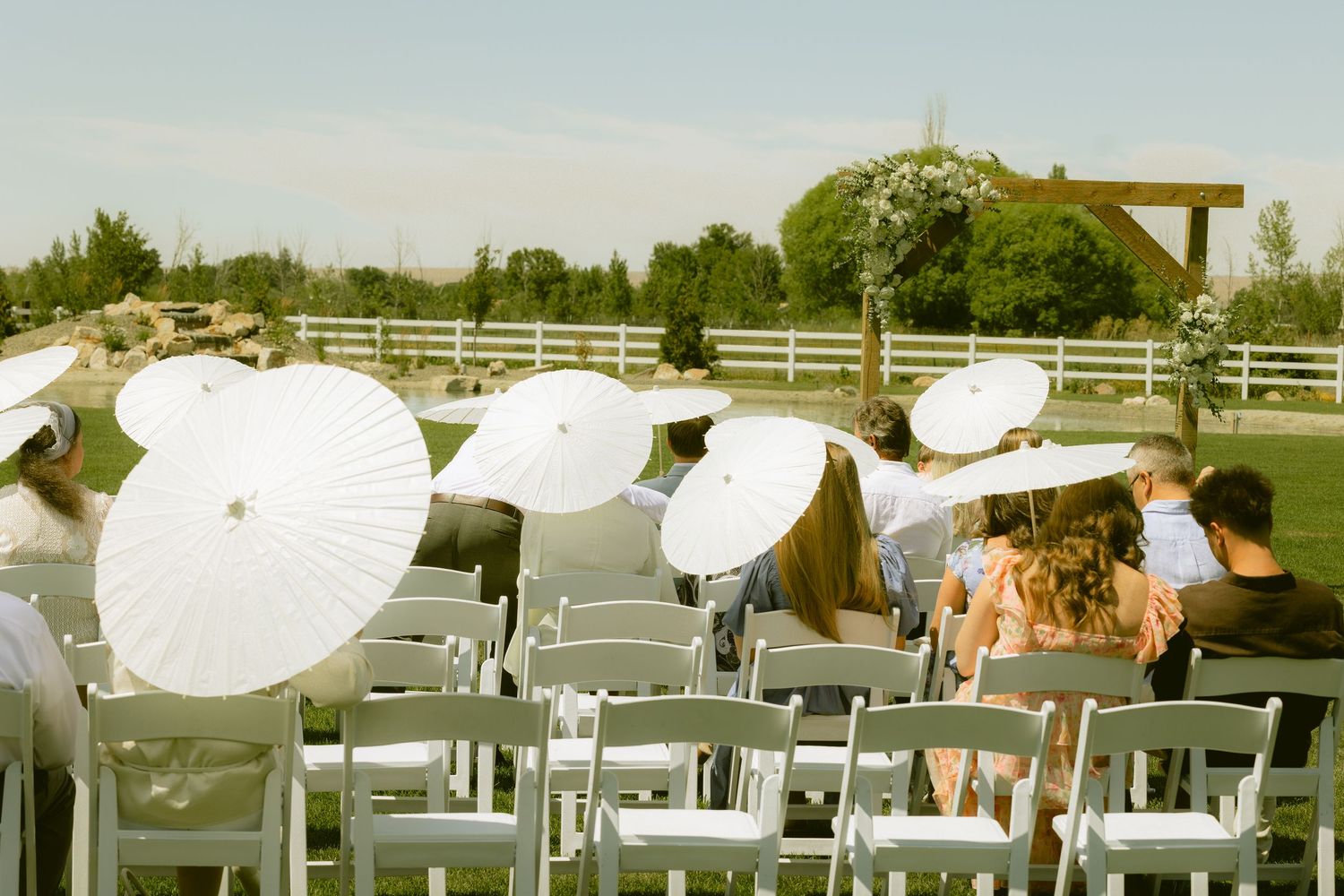 Wedding guests holding white parasols seated in white chairs at an outdoor ceremony with a wooden arch backdrop.