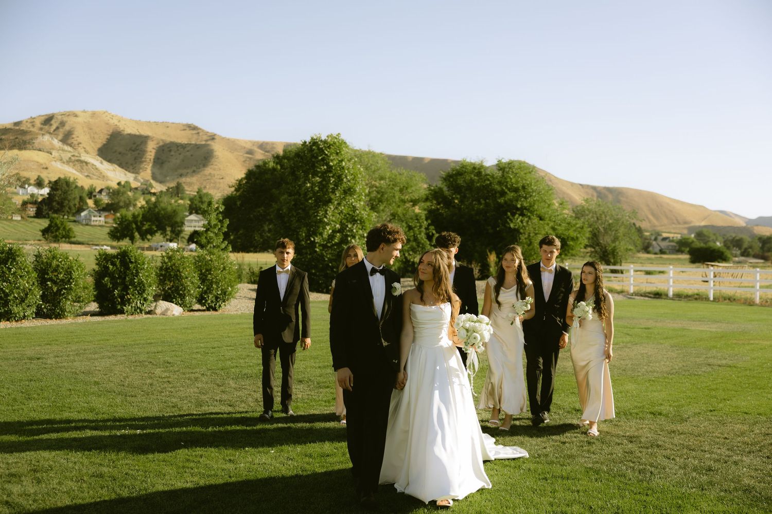 Bride and groom walking with wedding party across green lawn with scenic mountain backdrop.