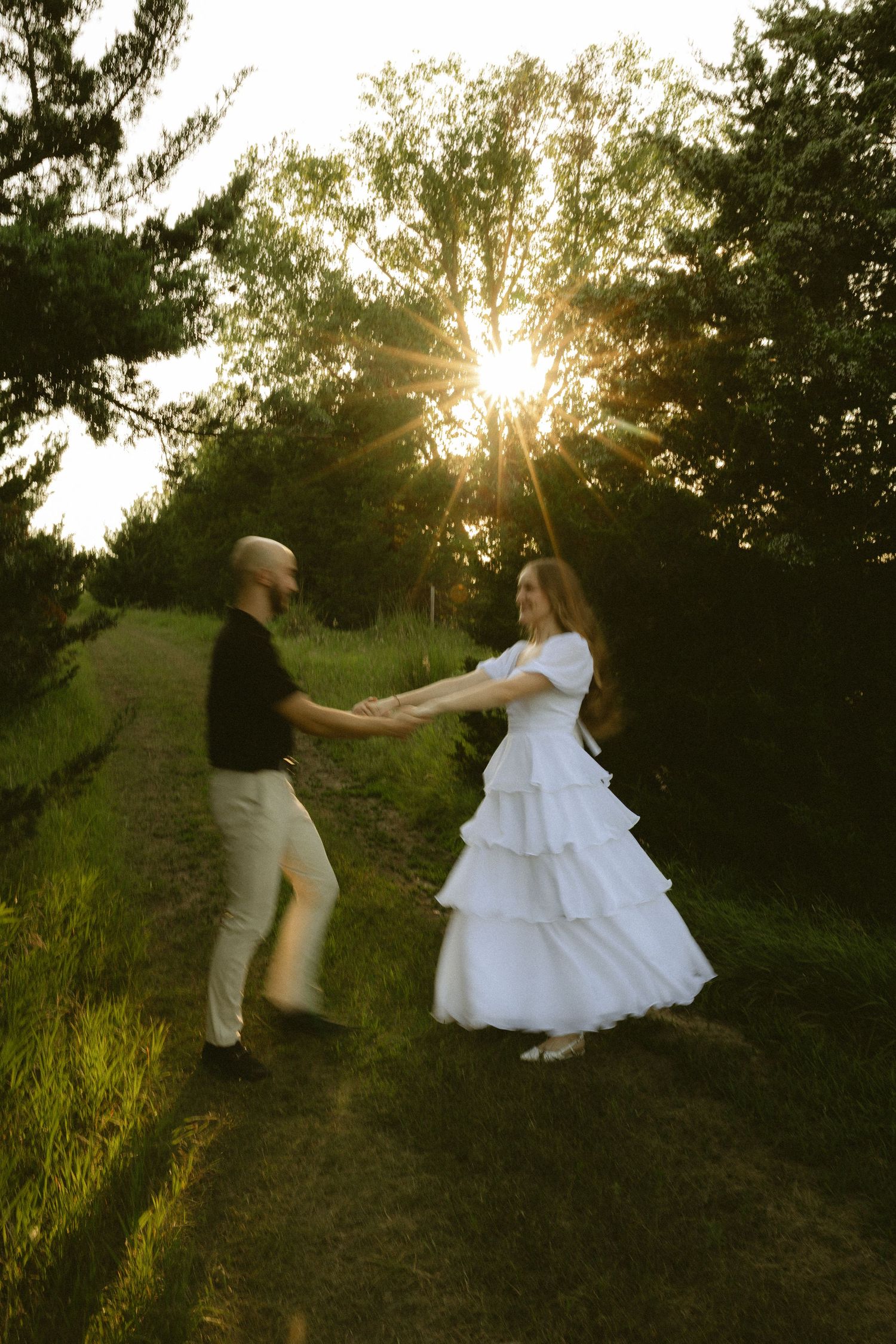 Couple dancing in a sunlit forest path, bride in white tiered dress holding hands with groom in black shirt at sunset.
