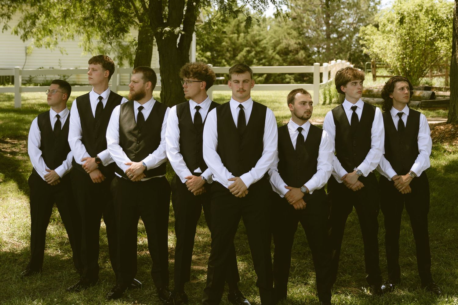Seven groomsmen in matching black vests, white shirts, and black ties stand in a line outdoors on a sunny day.