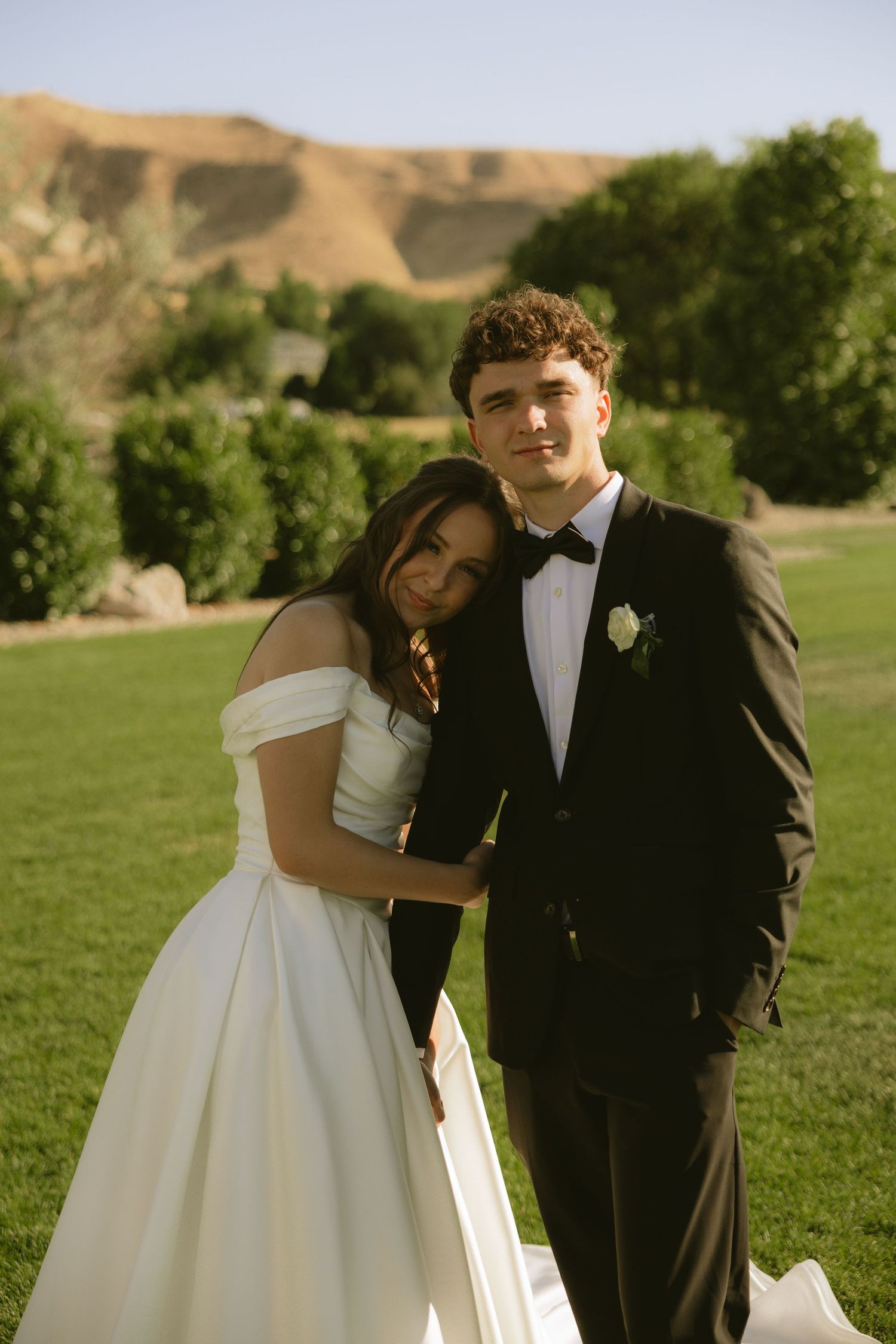 Bride in off-shoulder white gown leans on groom in black tuxedo outdoors on green lawn with sandy hills behind.