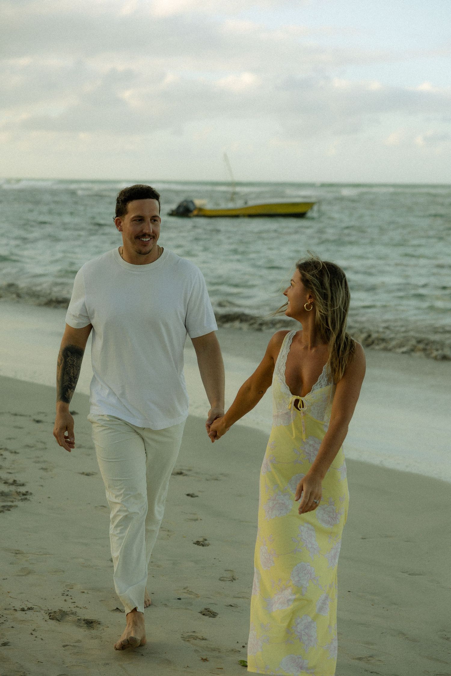 A couple walks hand in hand along a beach at sunset with a yellow boat visible in the ocean behind them.