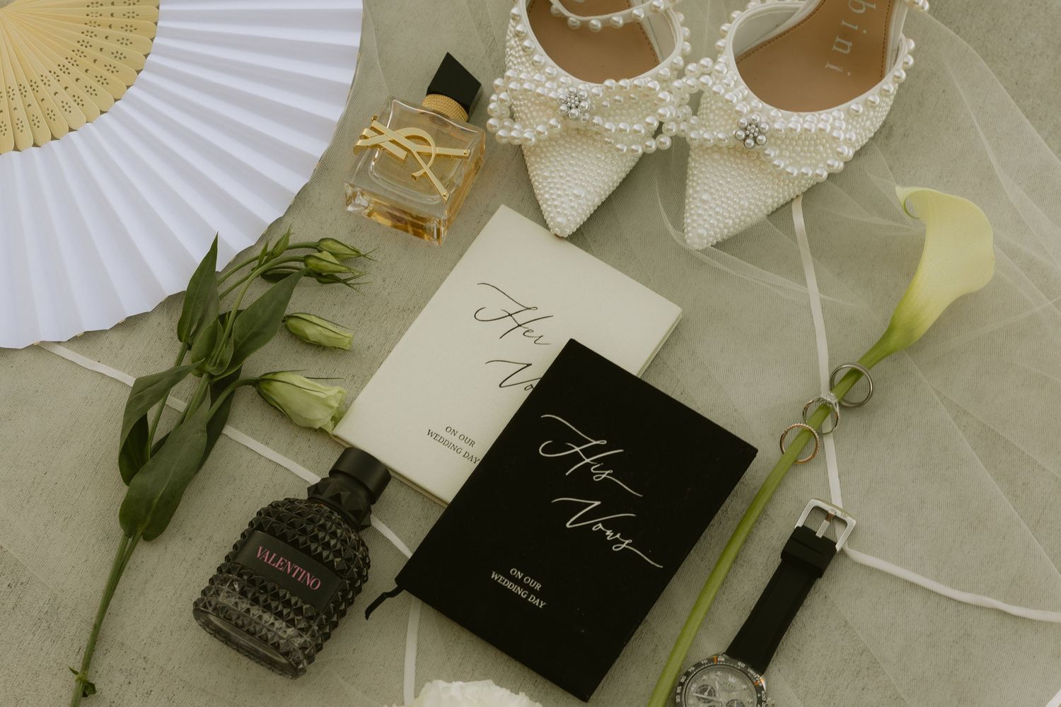 Elegant wedding flat lay with pearl heels, white fan, calla lily, vow books, perfume, and watch on tile.