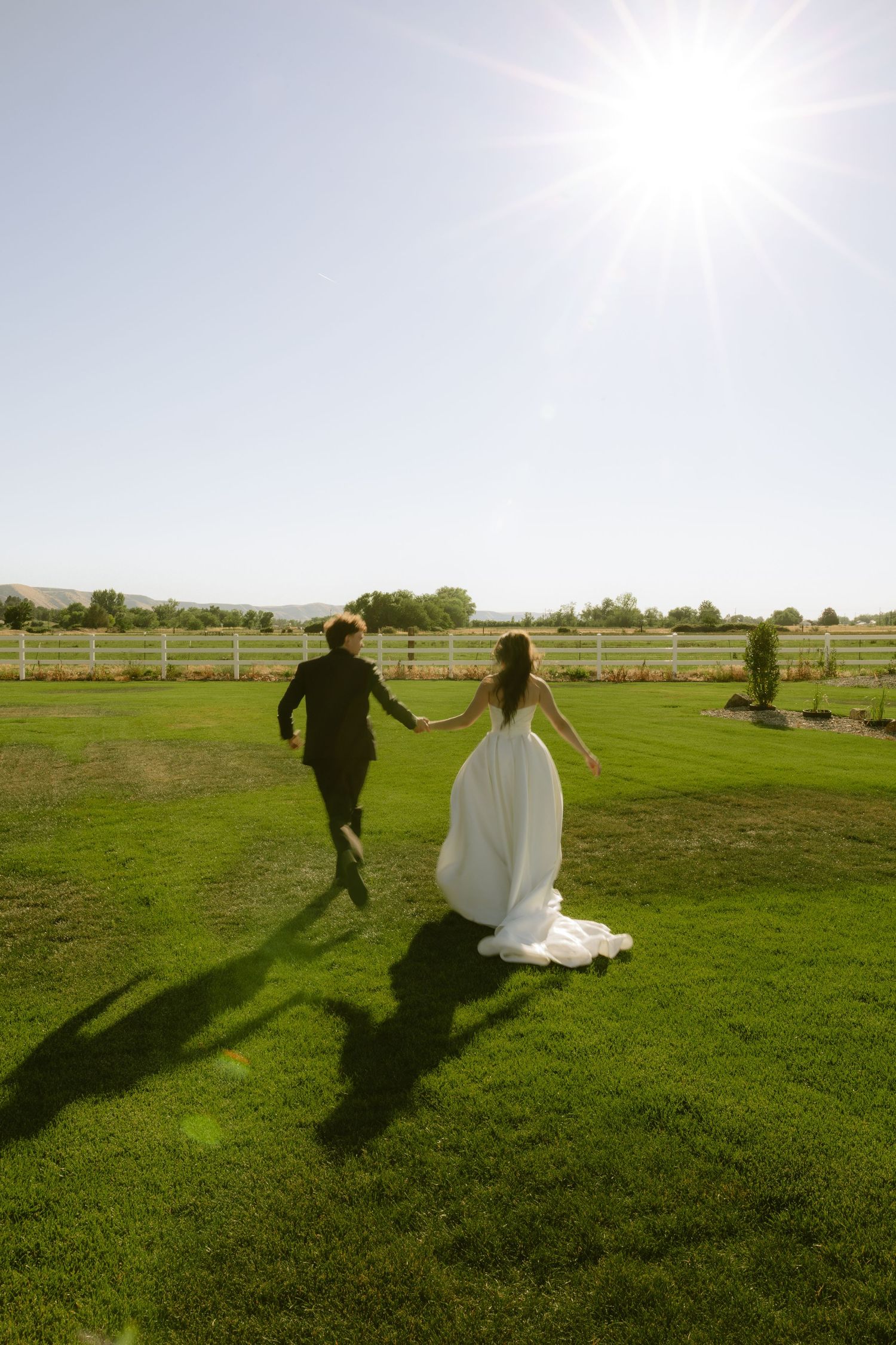 Bride and groom holding hands running across lush green lawn at sunny outdoor wedding venue with white fence.