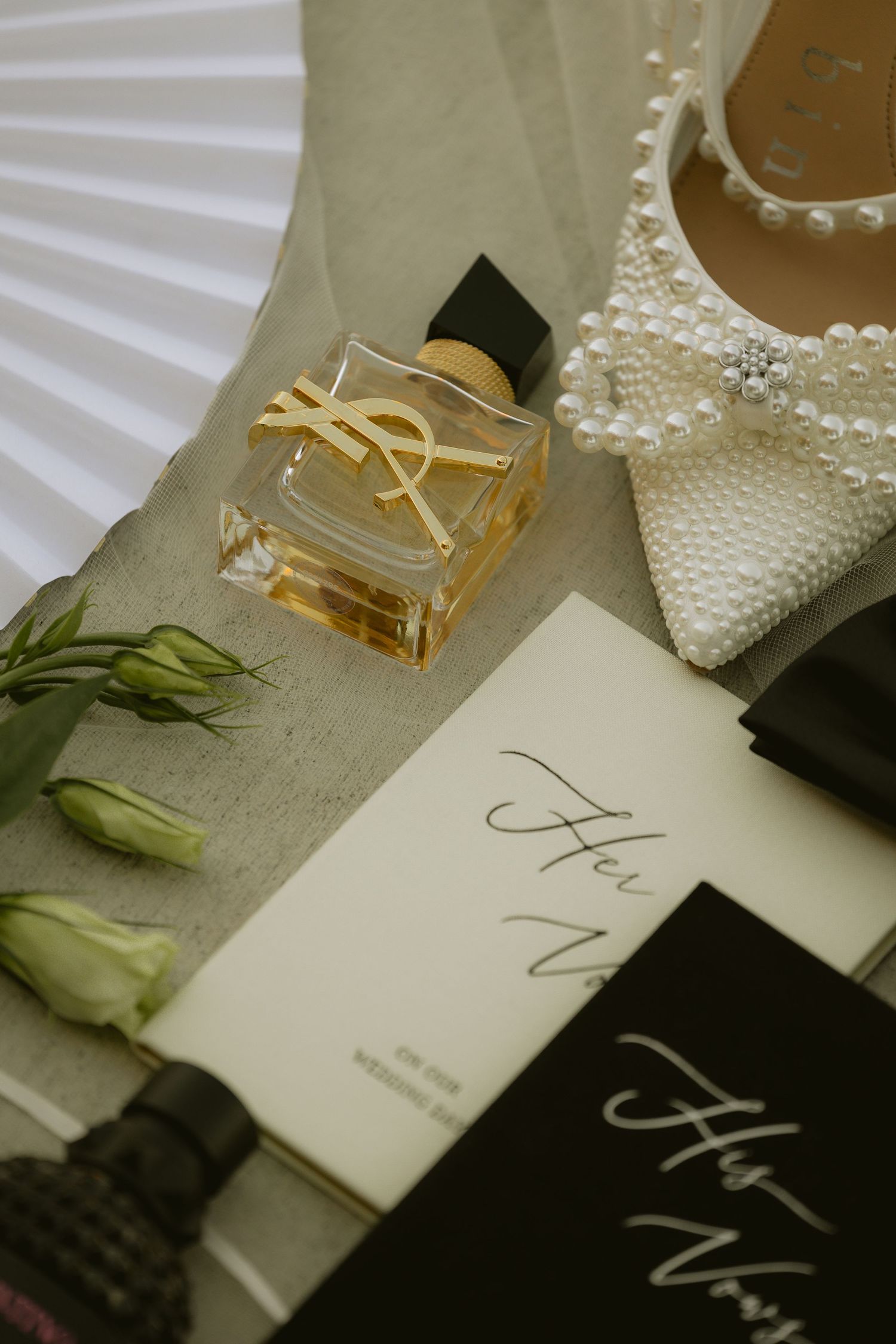 Wedding flat lay featuring YSL perfume, pearl heels, vow books, and white flowers on elegant neutral background.