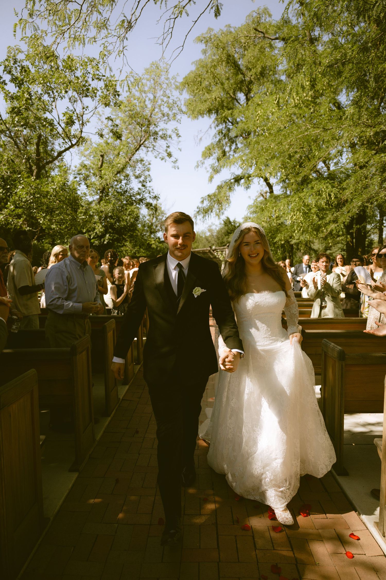 Newlywed couple walks down outdoor aisle in wedding attire, bride in white gown with red shoes, guests cheering around them.