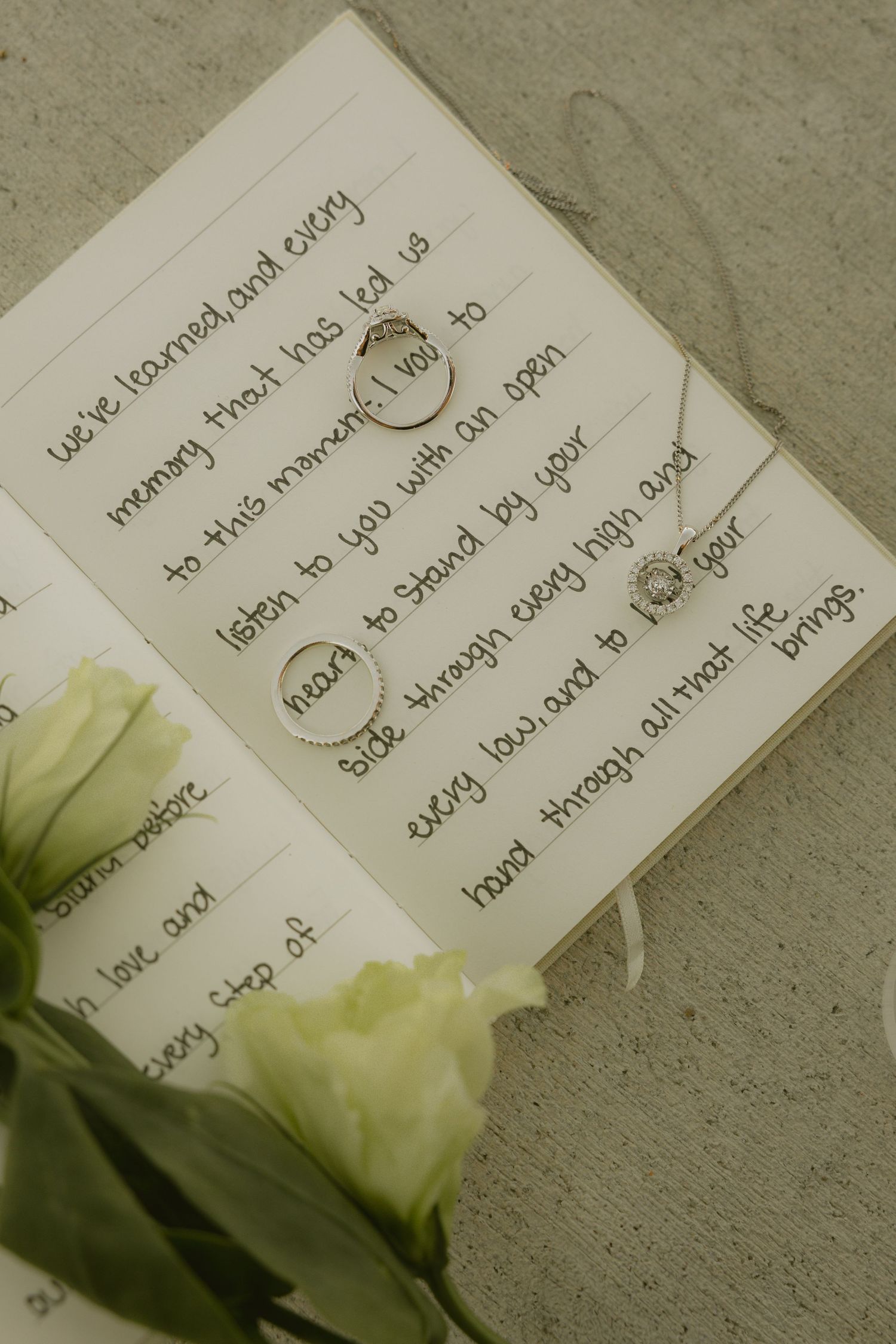 Wedding rings and necklace rest on open vow book with handwritten vows beside white roses.