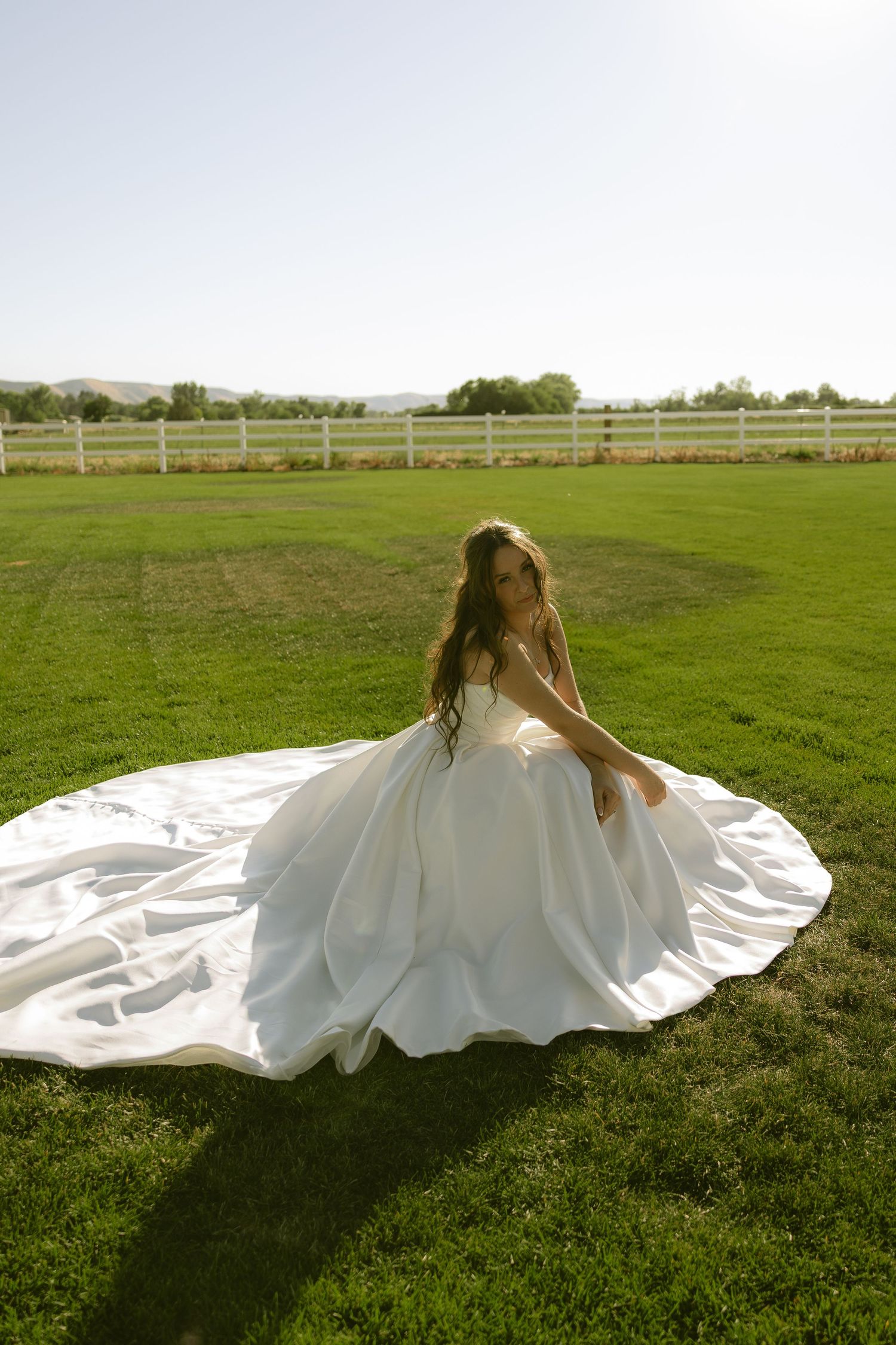Bride in a stunning white ball gown with long train sitting on green grass outdoors.