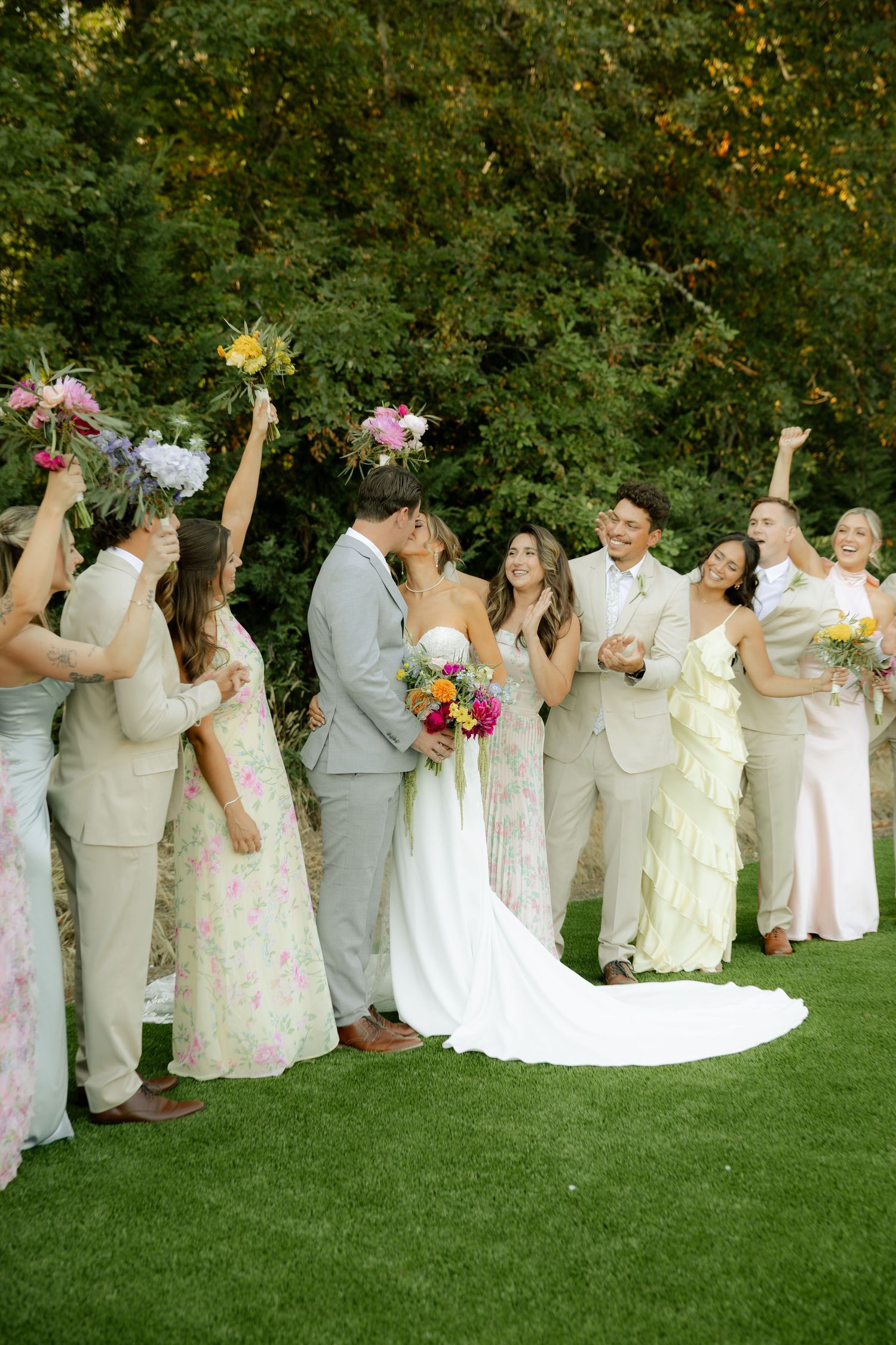Wedding party in floral dresses and gray suits poses on green lawn in playful formation.