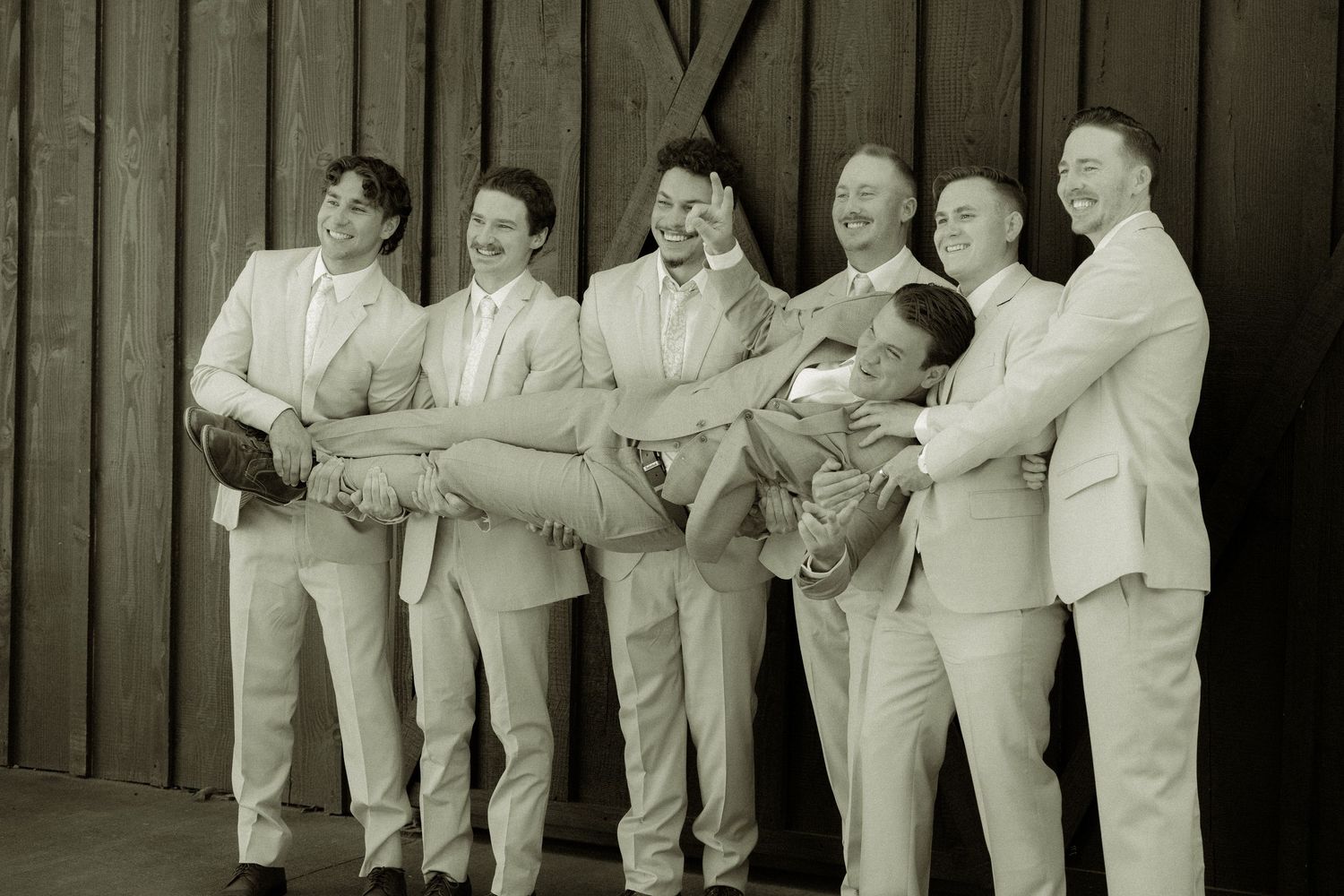 Groomsmen in light suits pose against dark wooden wall in monochrome photo.