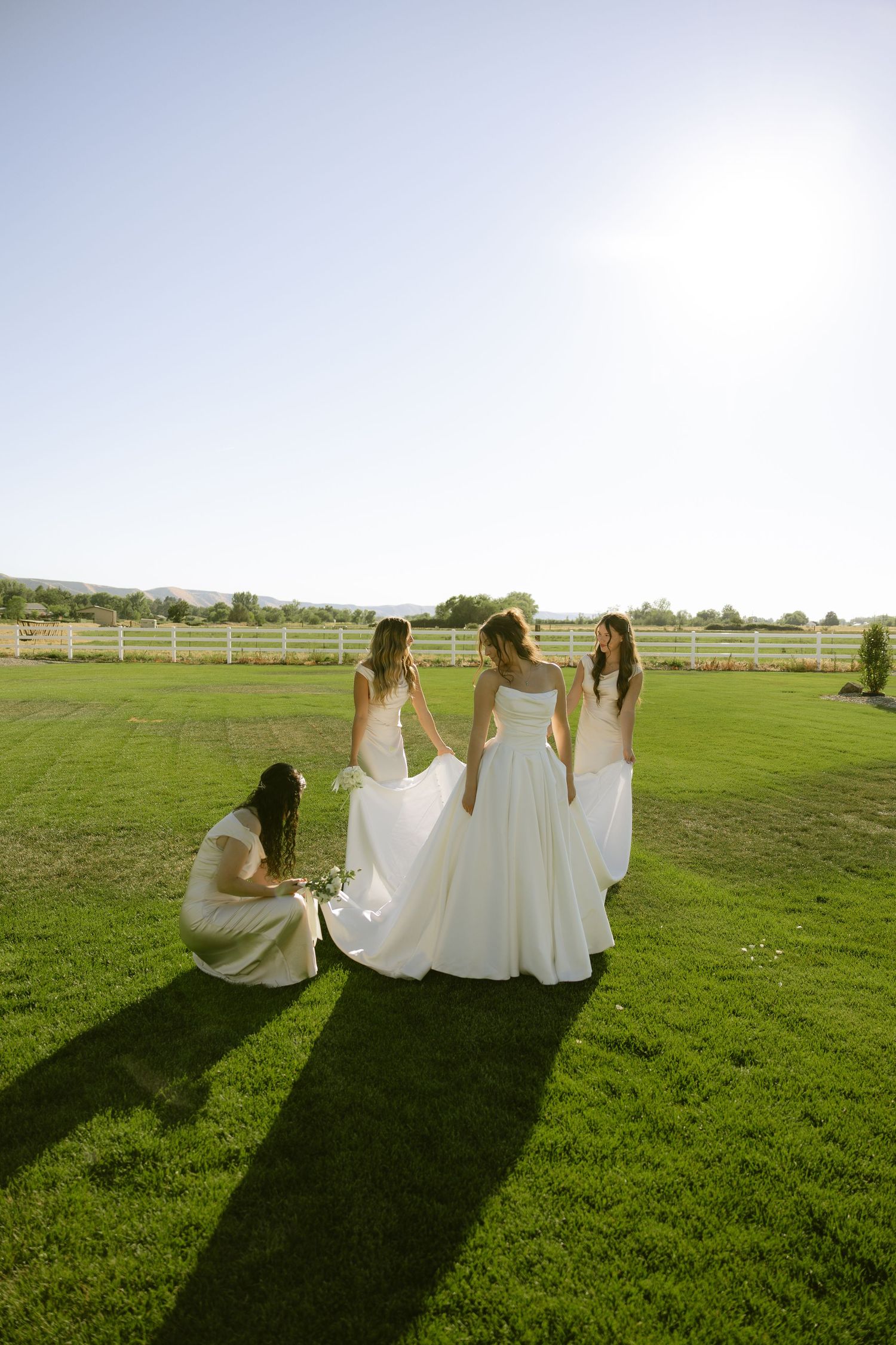 Bride stands with three bridesmaids in white dresses on sunny green lawn near white fence.