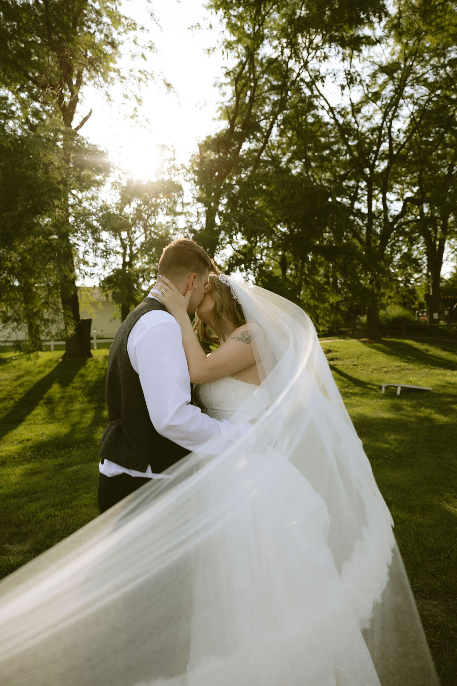 Bride and groom share a romantic kiss outdoors, with her flowing veil billowing in the breeze against a sunlit green backdrop.
