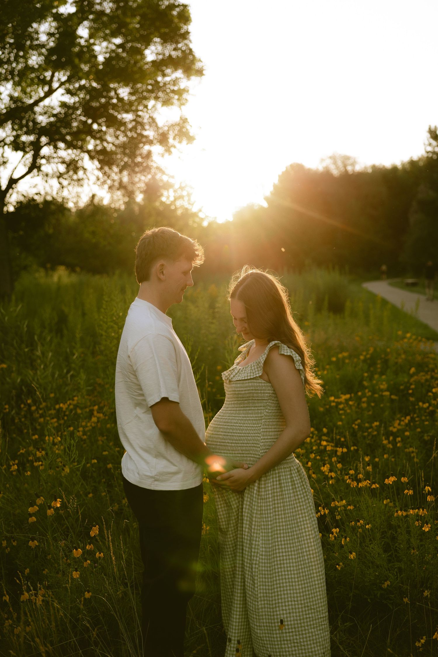 Couple embracing in golden sunset field, pregnant woman in green dress with partner in white tee.