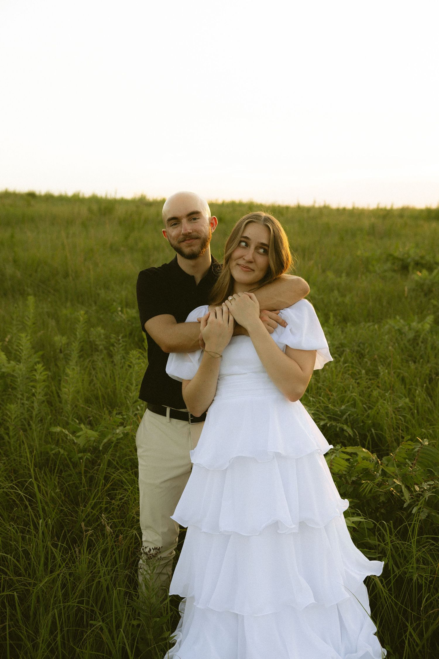 A bald man in black embraces a woman in a white tiered dress in a sunlit green field at golden hour.