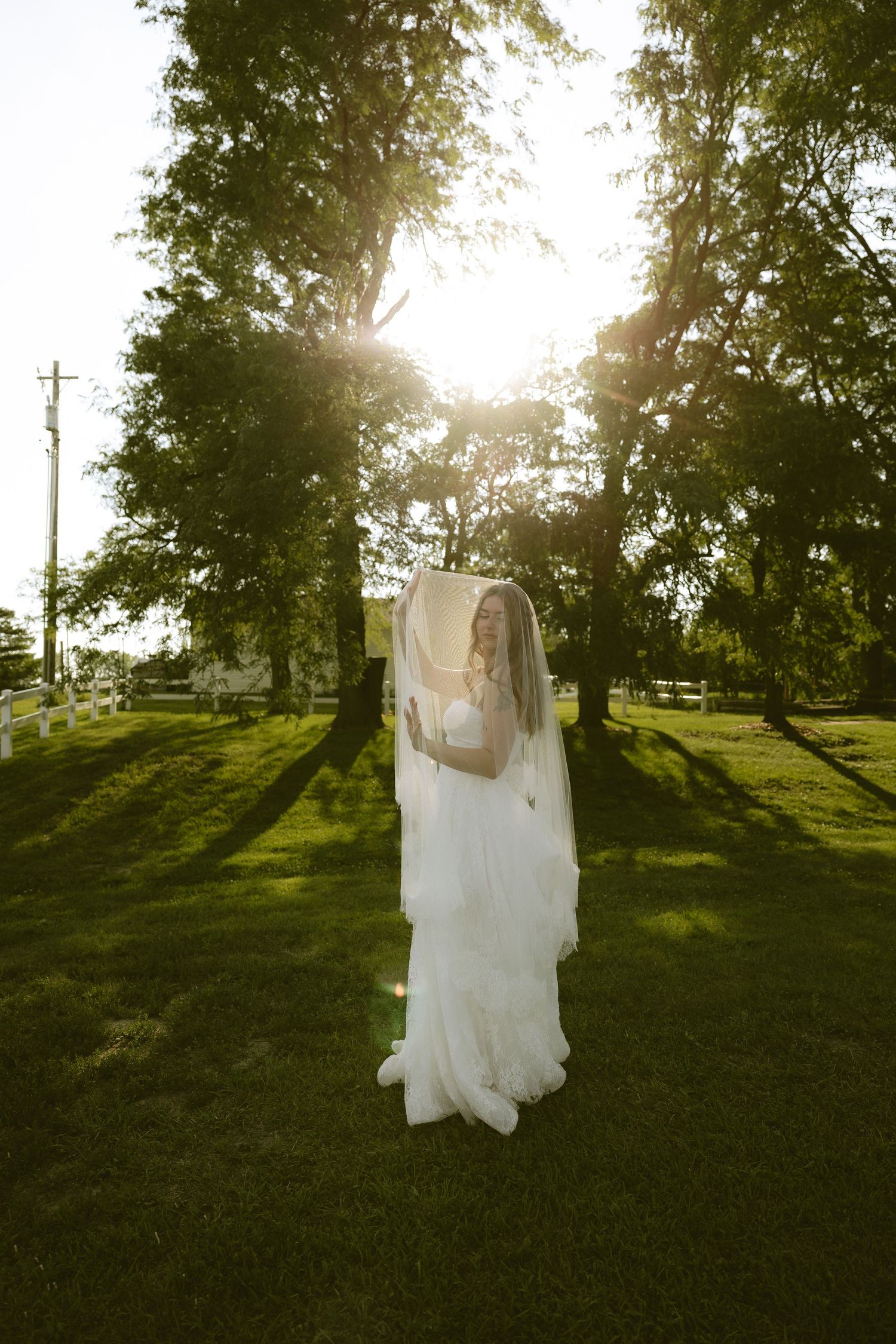 Bride in white gown and veil stands on lush green lawn with sunlight filtering through trees.