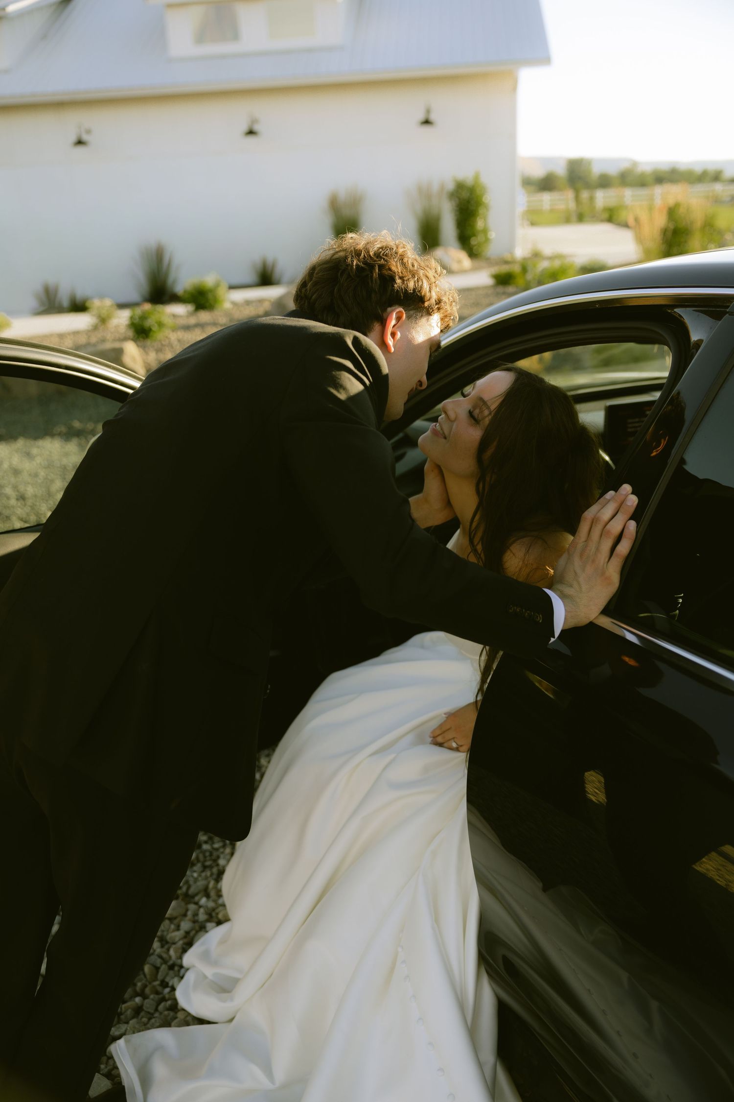 Groom in black suit leaning into car window to kiss bride in white wedding dress on their wedding day.