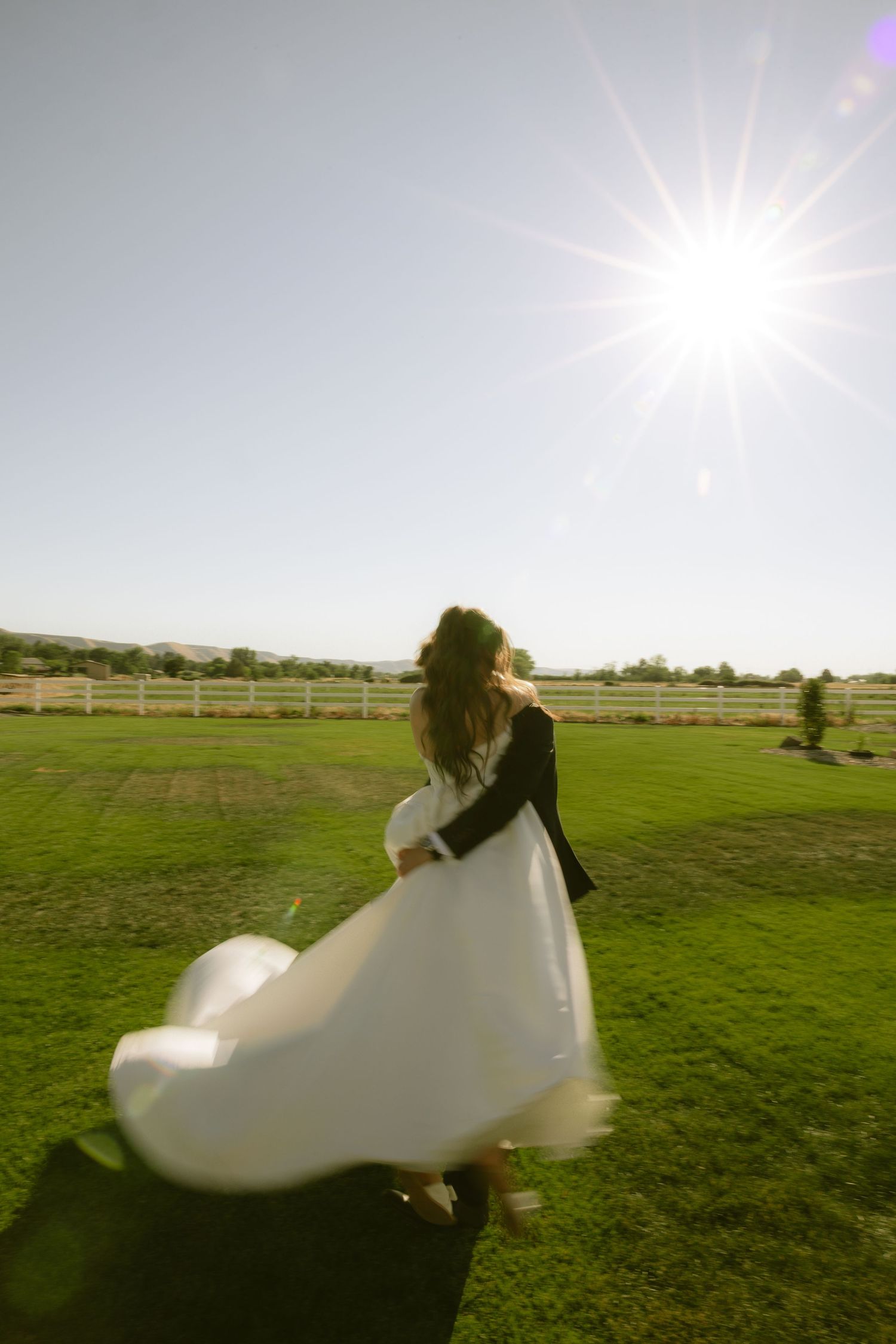 Groom twirls bride in flowing white dress on green farm lawn under bright sunburst sky.