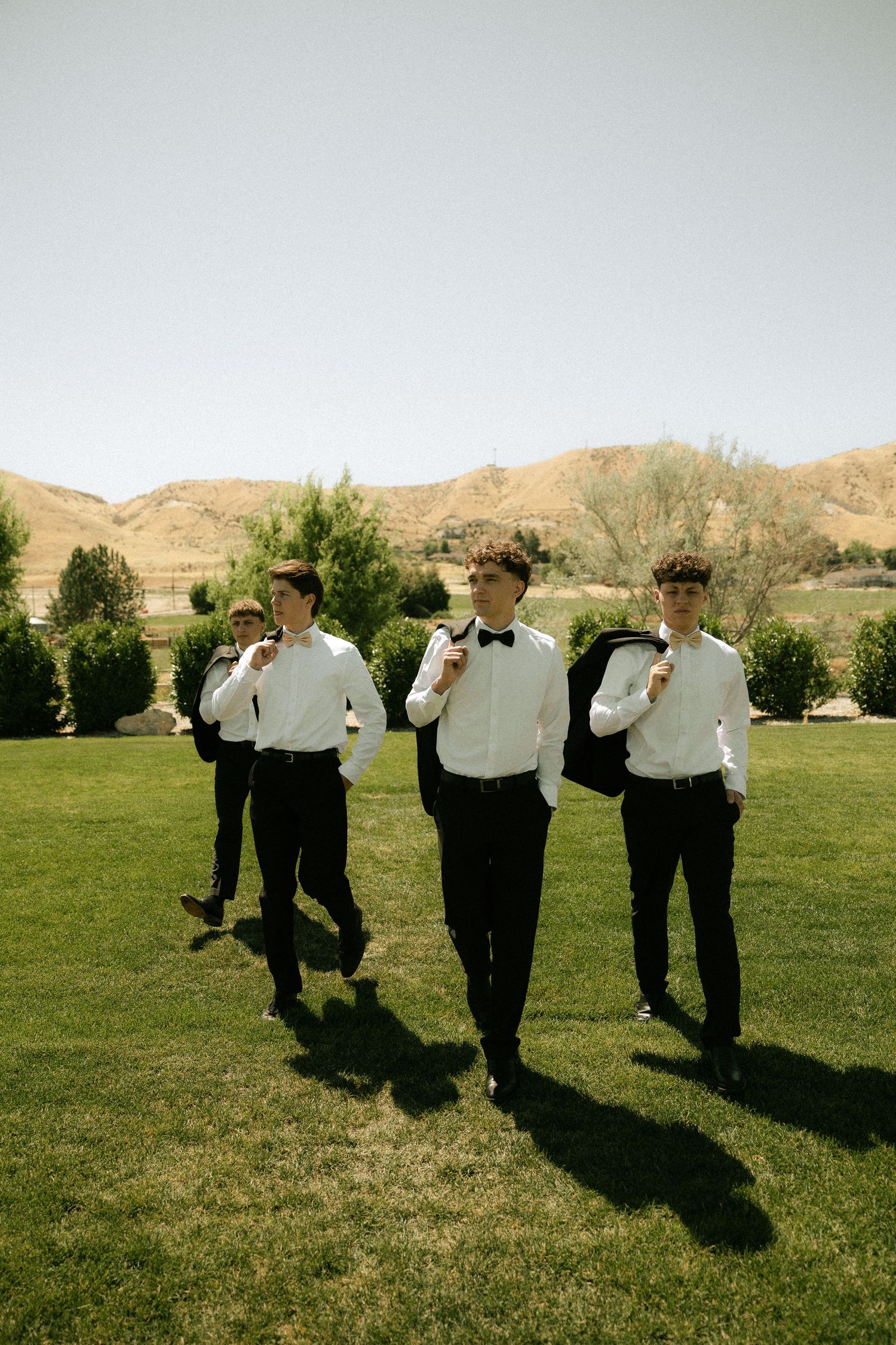 Four groomsmen in white shirts and black suits walking across green lawn with mountain backdrop.