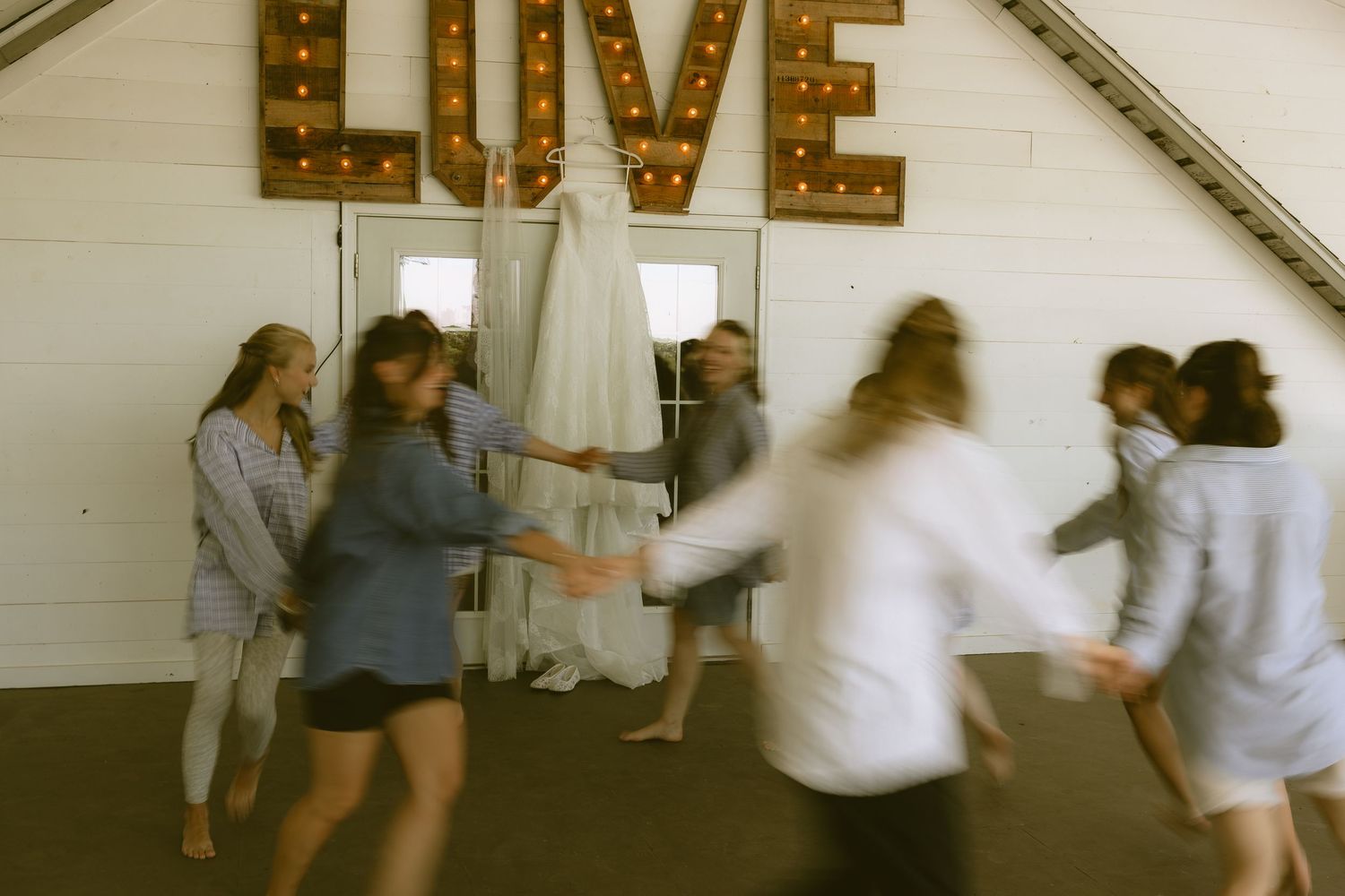 Bridesmaids in casual attire dance and celebrate around a wedding dress beneath a marquee LOVE sign in a barn.