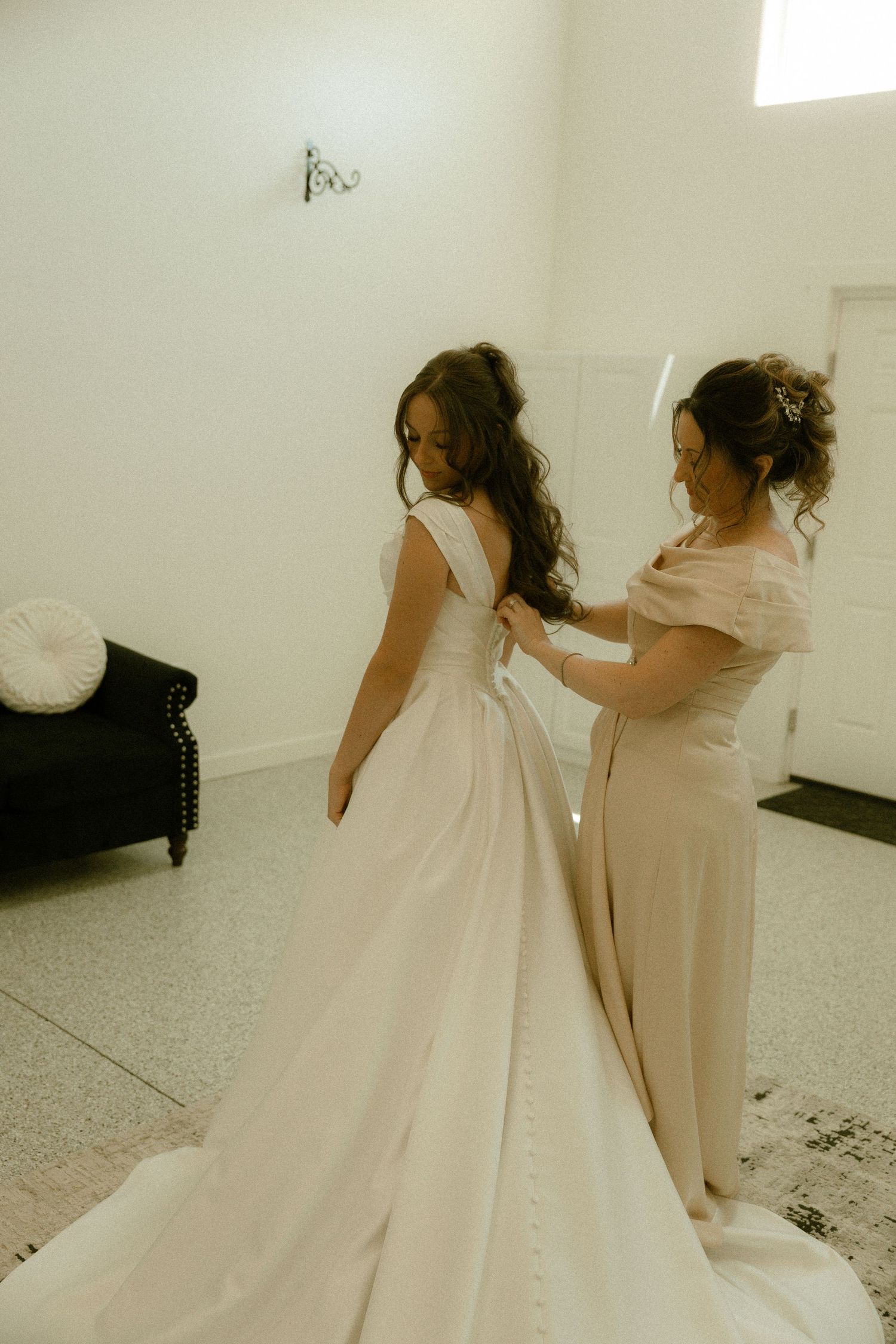 Bridesmaid helping bride lace up the back of her elegant white wedding gown indoors.