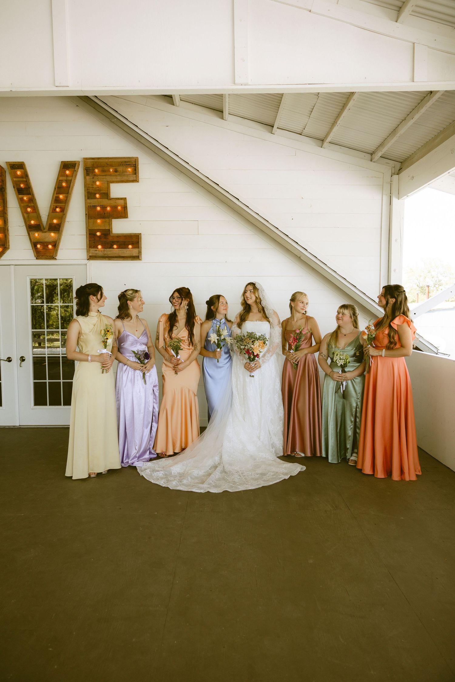 Bride with bridesmaids in colorful mismatched gowns posing near marquee LOVE sign at wedding venue.