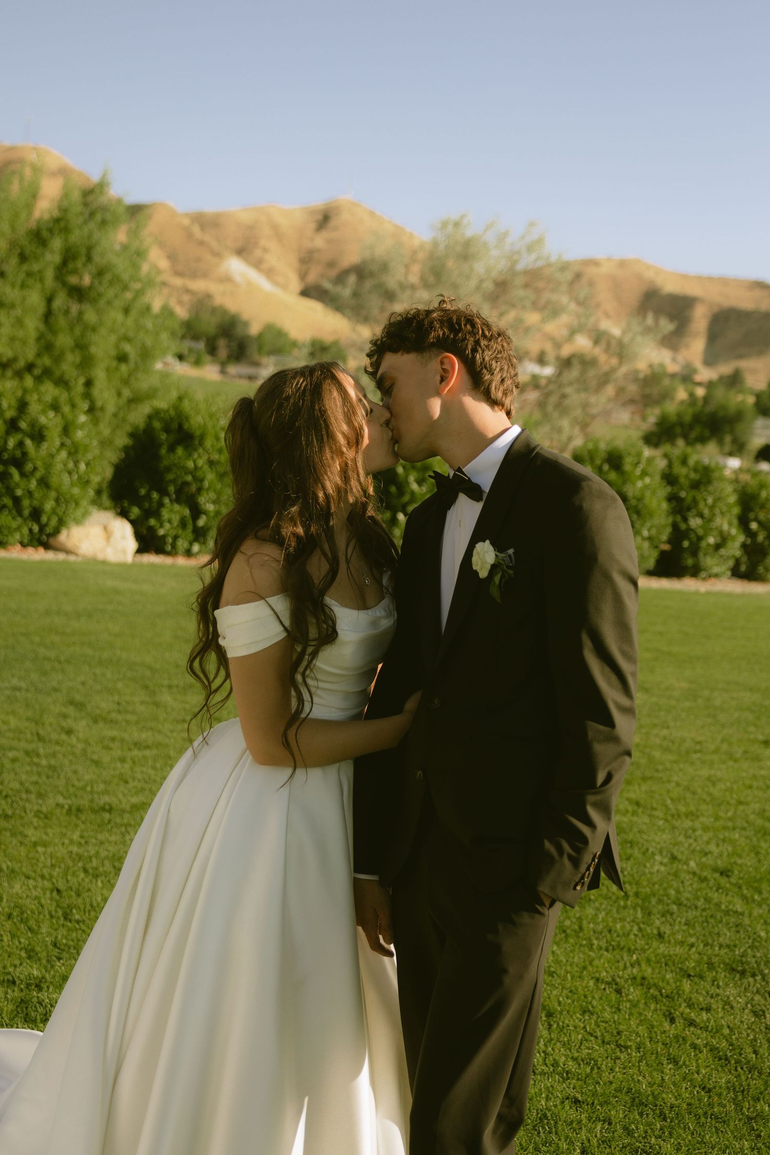 A bride and groom share a romantic kiss on a green lawn with scenic hills in the background on their wedding day.